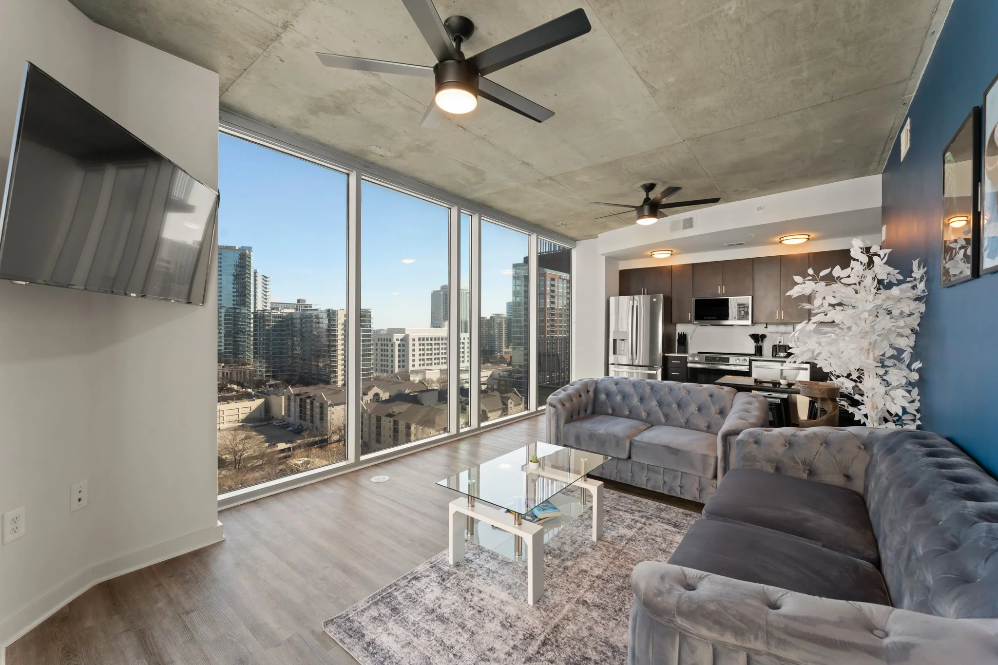 Modern apartment living room with large floor-to-ceiling windows, city skyline view, gray velvet sofas, glass coffee table, kitchen with stainless steel appliances, and decorative white tree.