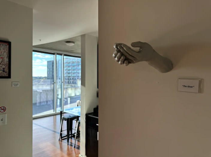A white sculpture of a hand placed on a wall with a sign that reads 'The Giver.' in a modern apartment interior, with large sliding glass doors leading to a balcony and city view.
