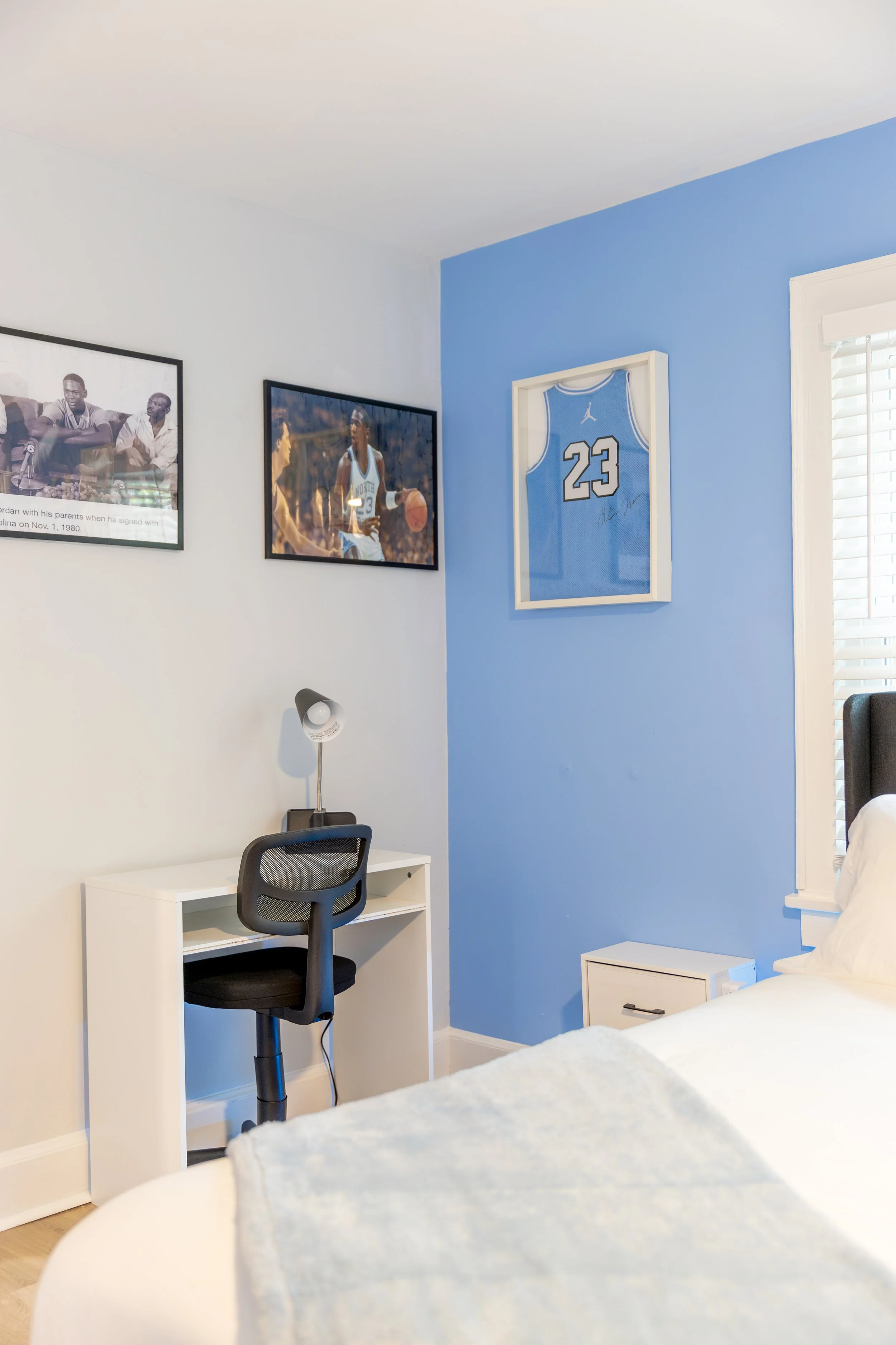 A bedroom with a white wall and blue accent wall, a framed basketball jersey on the blue wall, framed photos on the white wall, a small white desk with a black office chair, a white nightstand, a bed with white bedding, and window blinds.