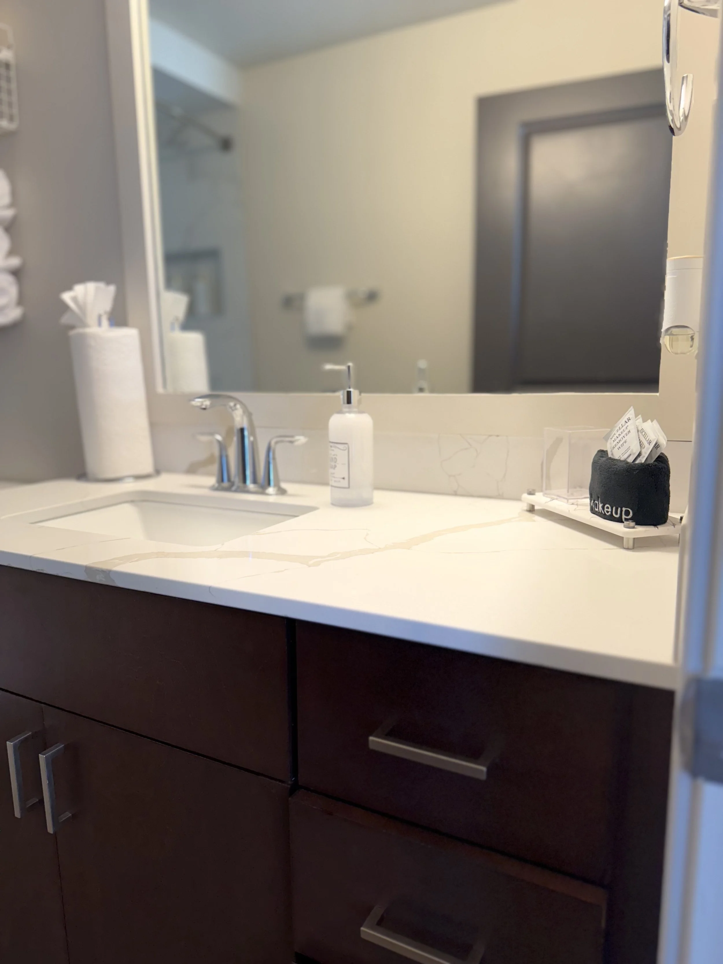 Bathroom vanity with a white marble countertop, a small sink, a chrome faucet, a soap dispenser, a roll of paper towels, and a black makeup brush holder with tissues, reflected in a large mirror. The dark wood cabinet has drawers with modern handles.