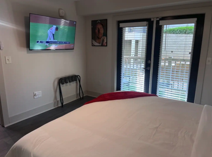 Hotel room with a bed, wall-mounted TV showing a baseball game, artwork on wall, and a glass door with blinds leading outside.