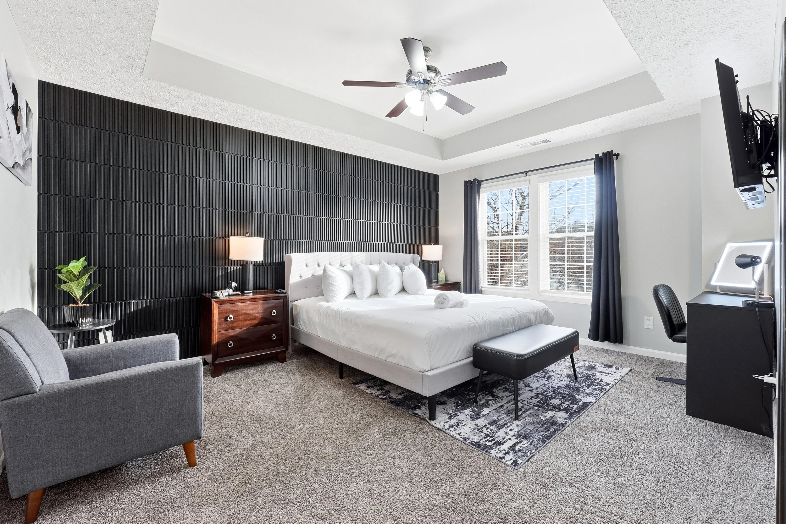 Bright living room with large windows, black sofas with blue pillows, a blue rug, and an open kitchen in the background.