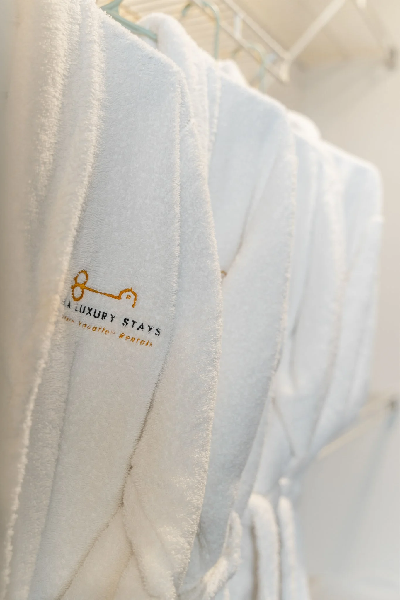 Close-up of white bathrobes hanging on a rack, with a small embroidered logo featuring a key and the words "Luxury Stays".