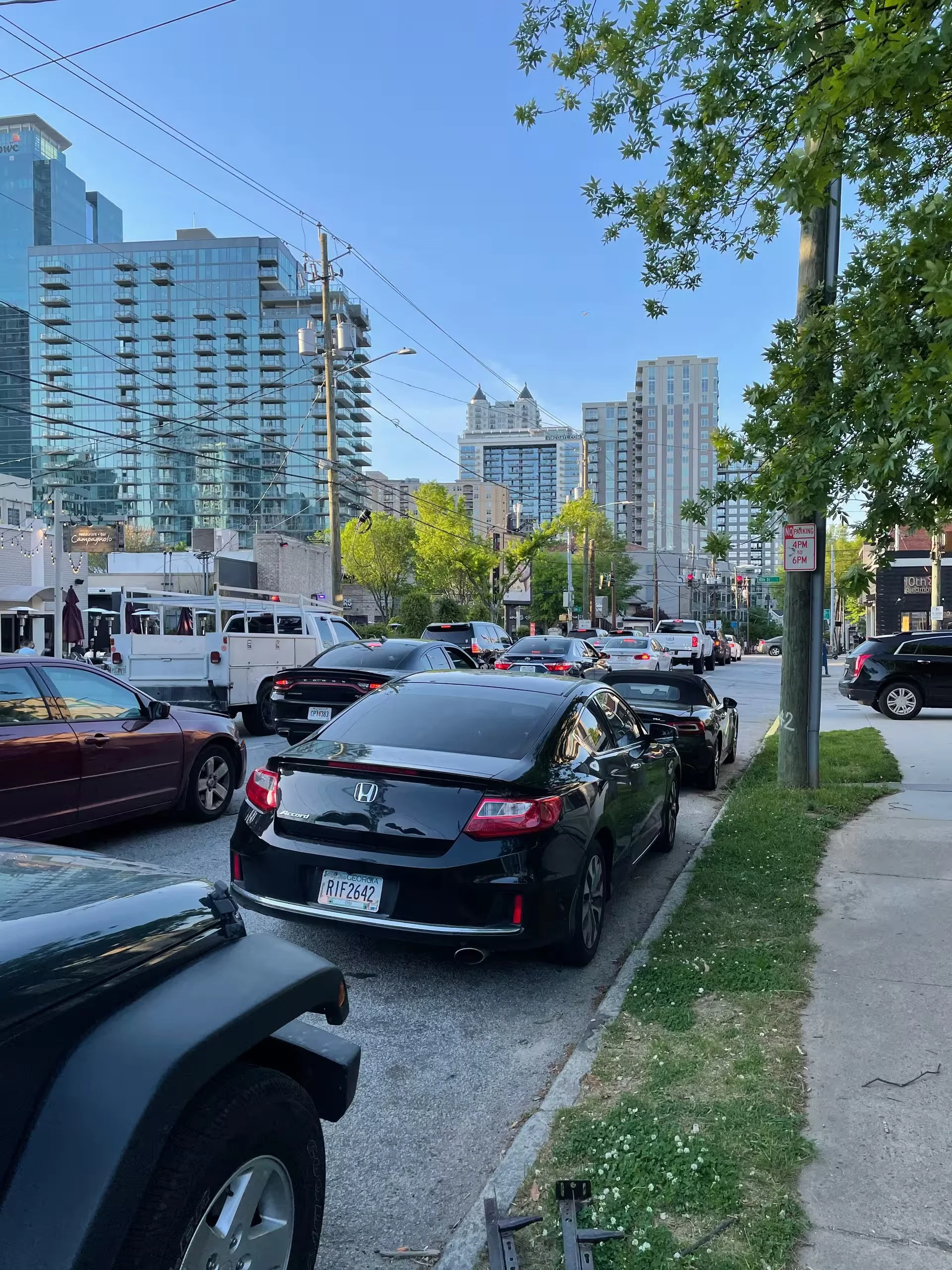A city street scene with parked cars along the sidewalk, tall modern buildings in the background, green trees, and power lines overhead on a clear day.