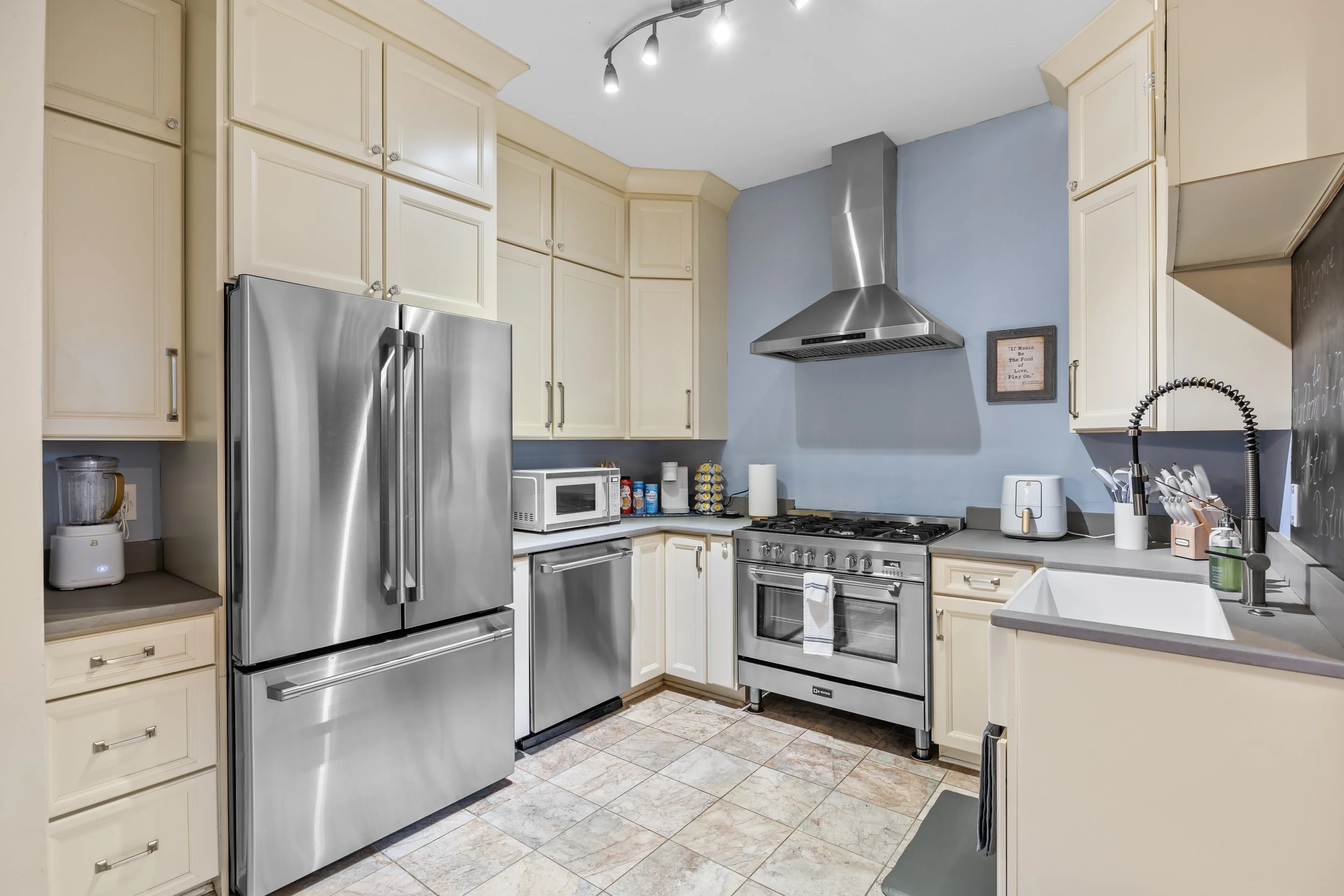 Kitchen with beige cabinets, stainless steel refrigerator, microwave, gas stove with range hood, and a blackboard with writing on the right wall.
