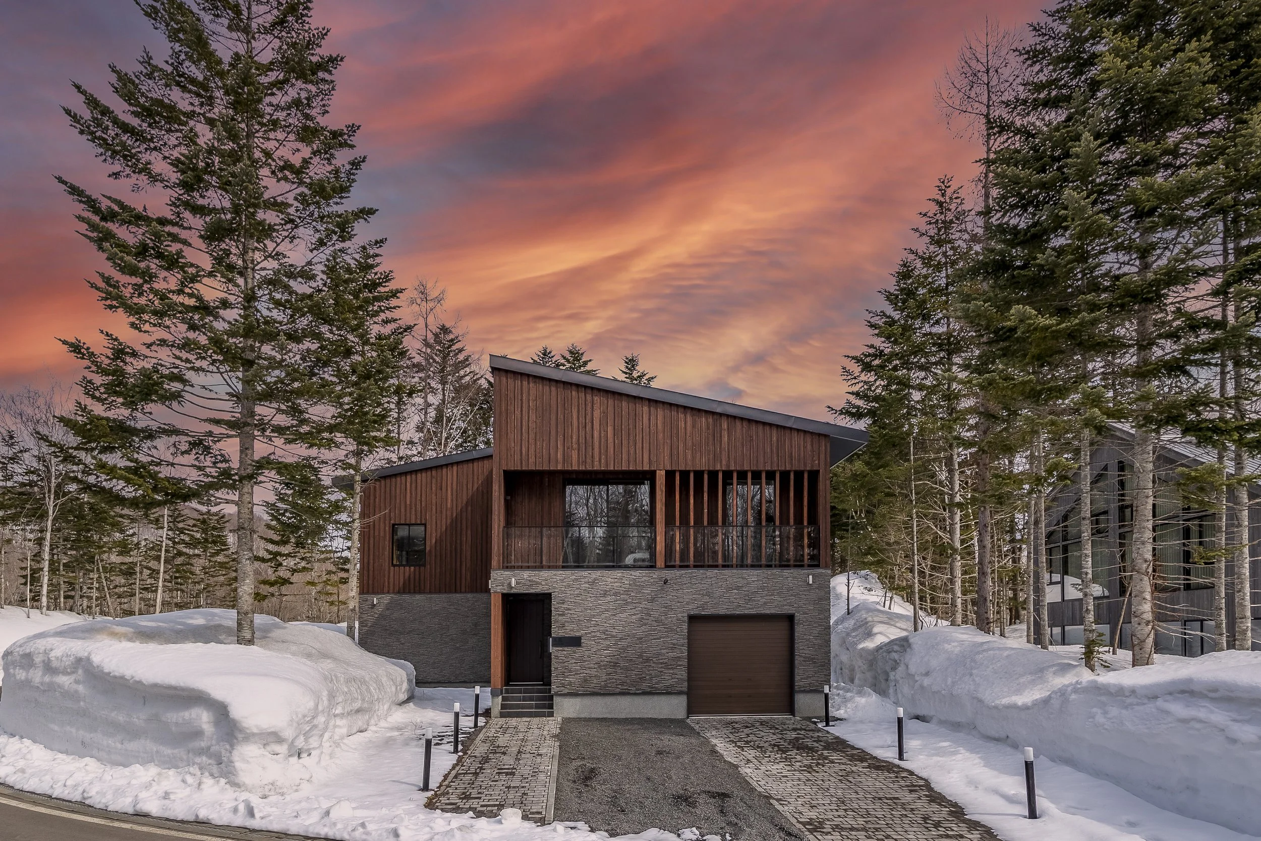 Modern wooden house with large windows in a snowy landscape at sunset, surrounded by tall pine trees and snow-covered ground.