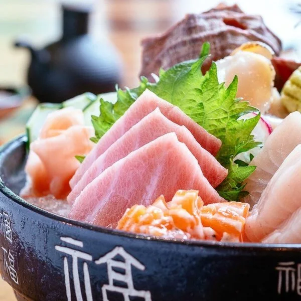 A black bowl with Japanese characters containing sliced pink ham, leafy green garnish, and various raw seafood such as scallops and fish, with a blurred background.