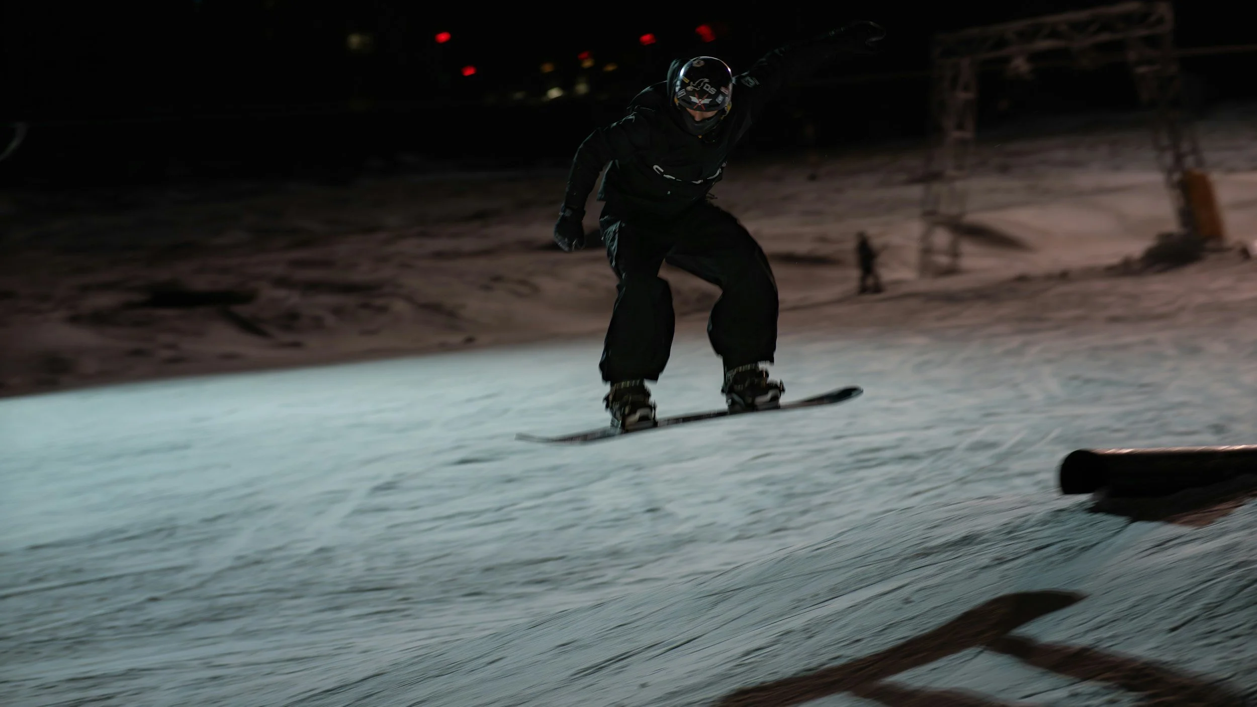 A person dressed in black snowboarding gear, including a helmet, snowboarding down a slope at night under artificial lights.