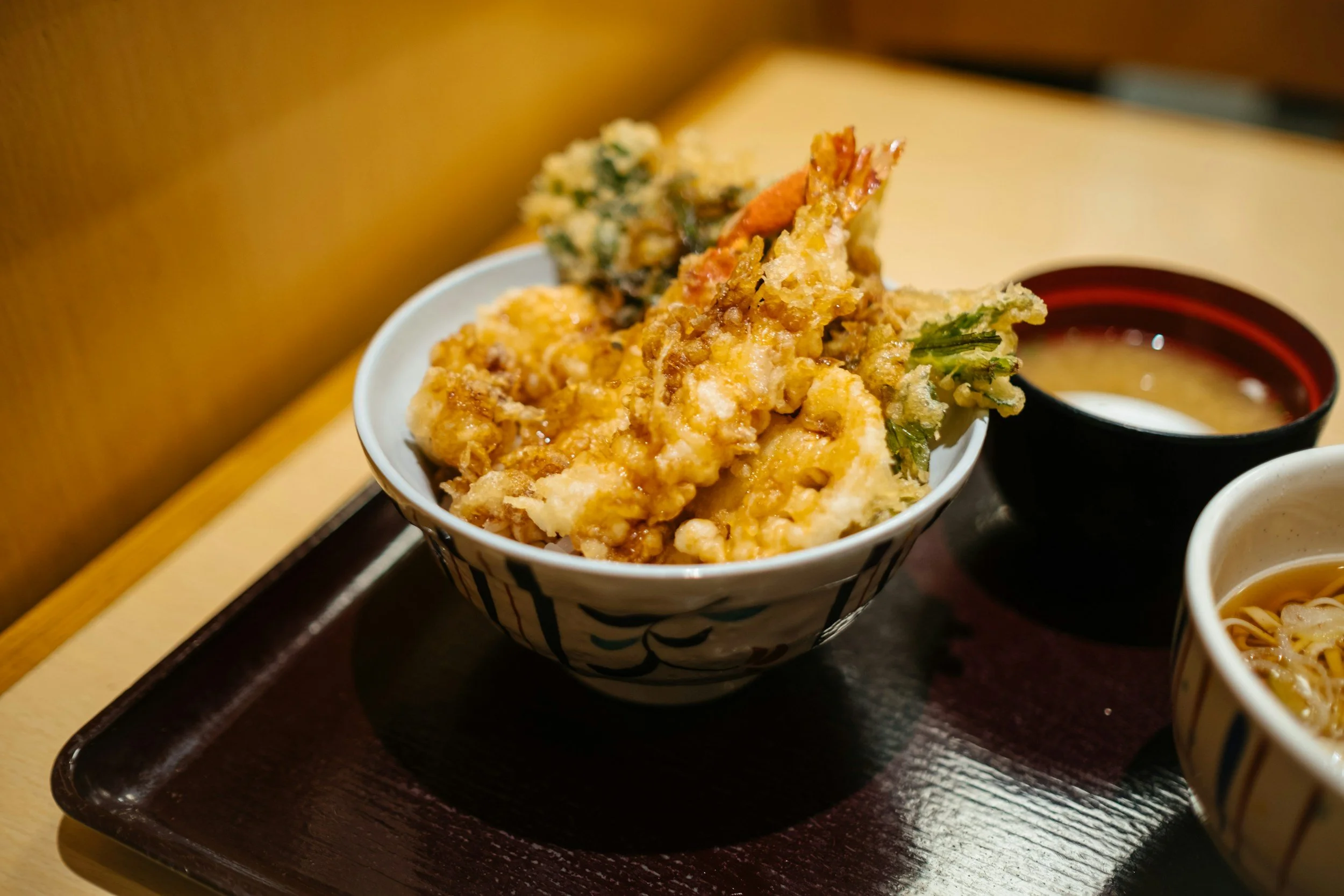 A bowl of tempura with mixed vegetables and shrimp, accompanied by dipping sauces on a wooden tray.