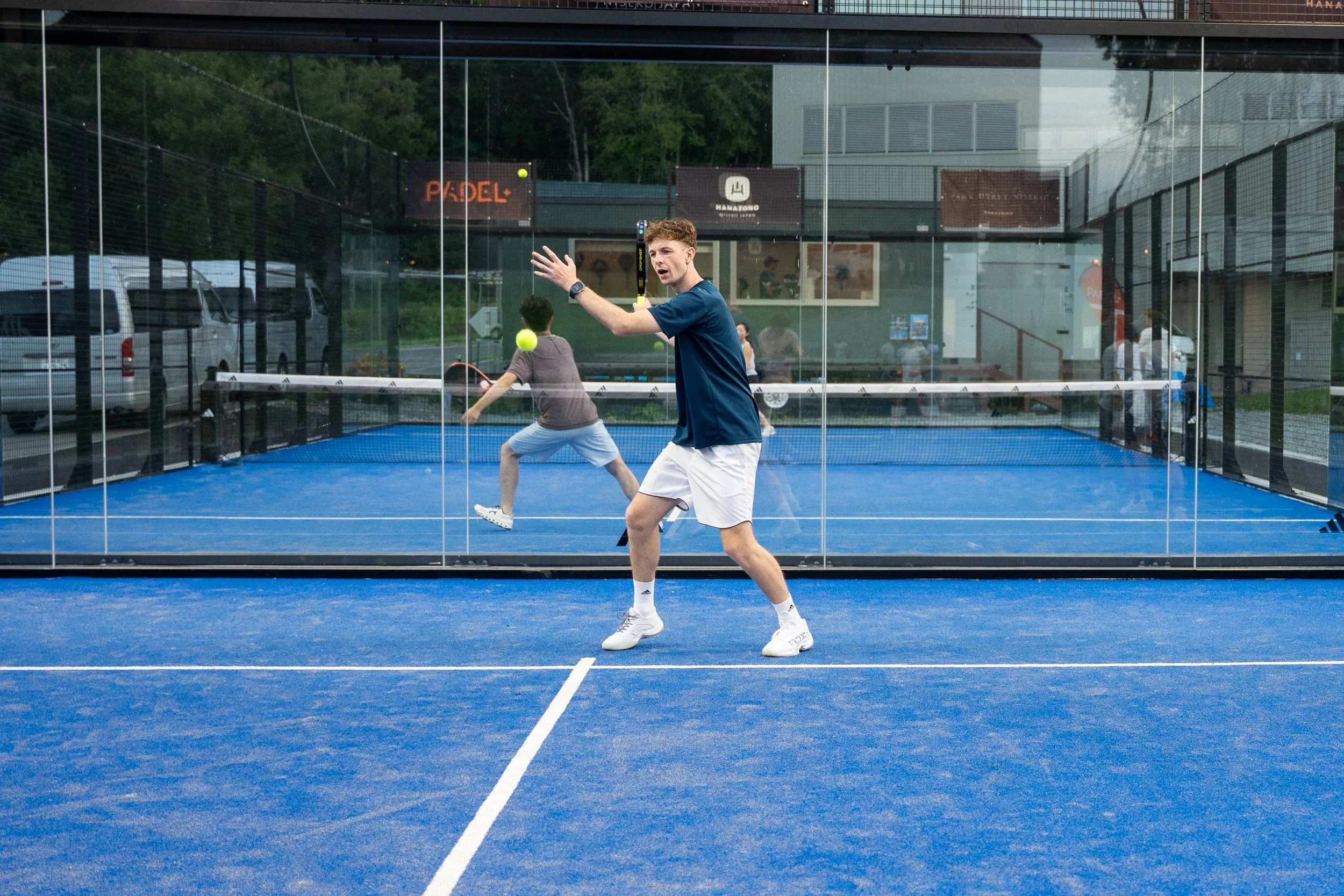 Two young men playing padel on a blue court enclosed by glass walls with a city street and parked cars visible outside.