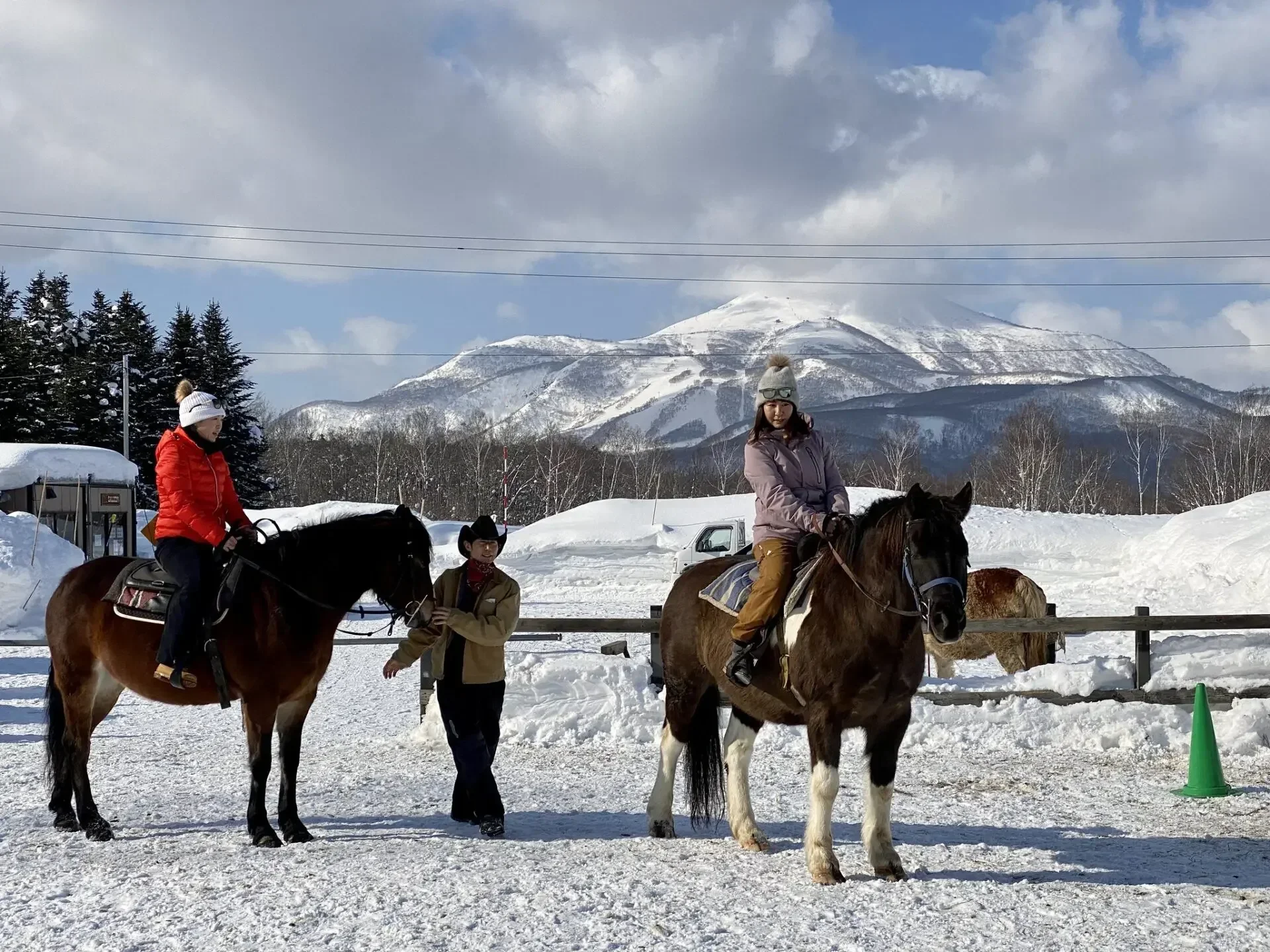 People riding horses in a snowy outdoor setting with mountains and pine trees in the background.