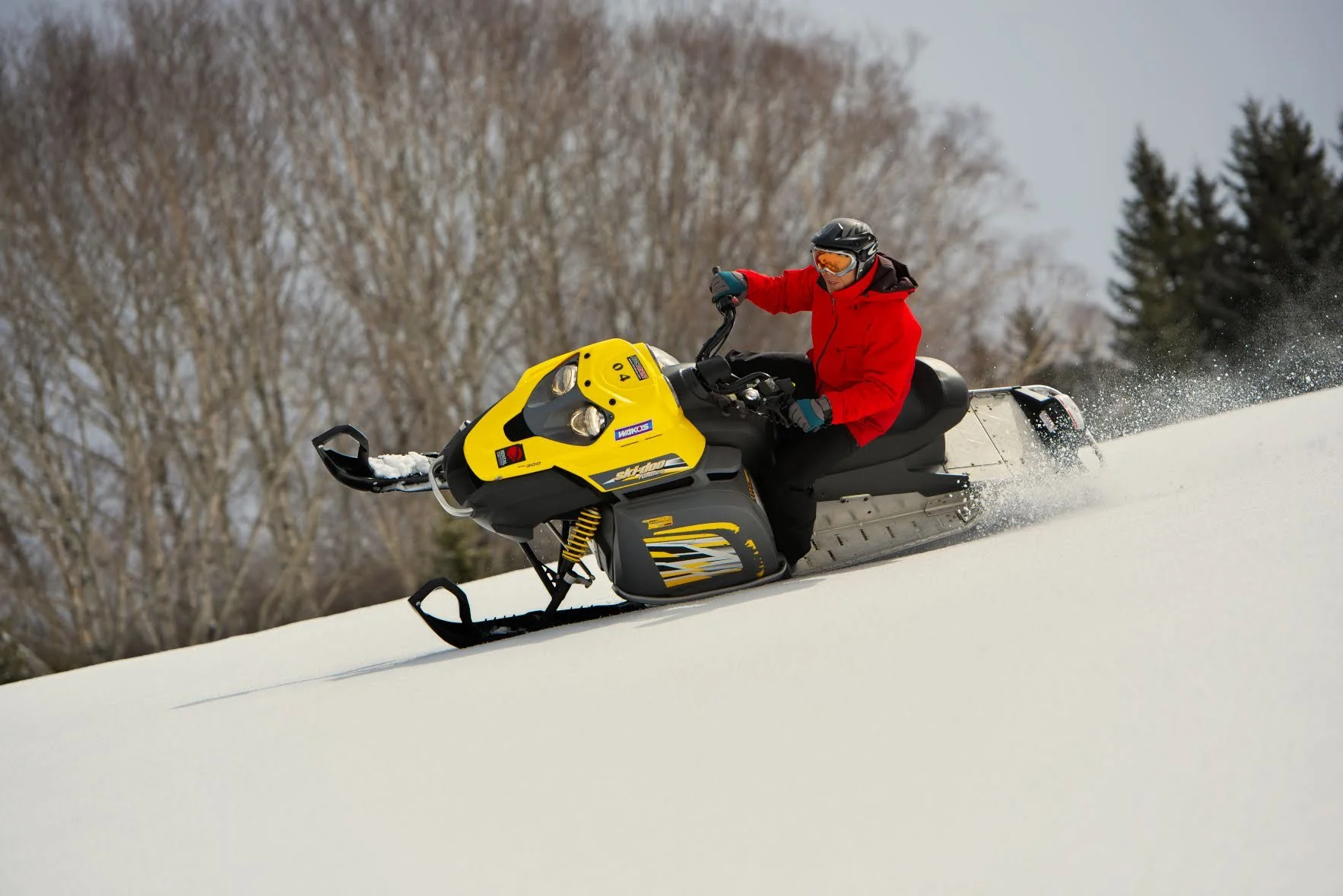 Person wearing a red jacket and goggles riding a yellow snowmobile on snow with snow-covered trees in the background.