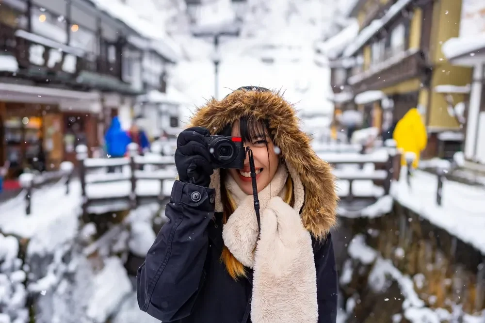 A woman in winter clothing taking a photo with a camera on a snowy street in a village or town.