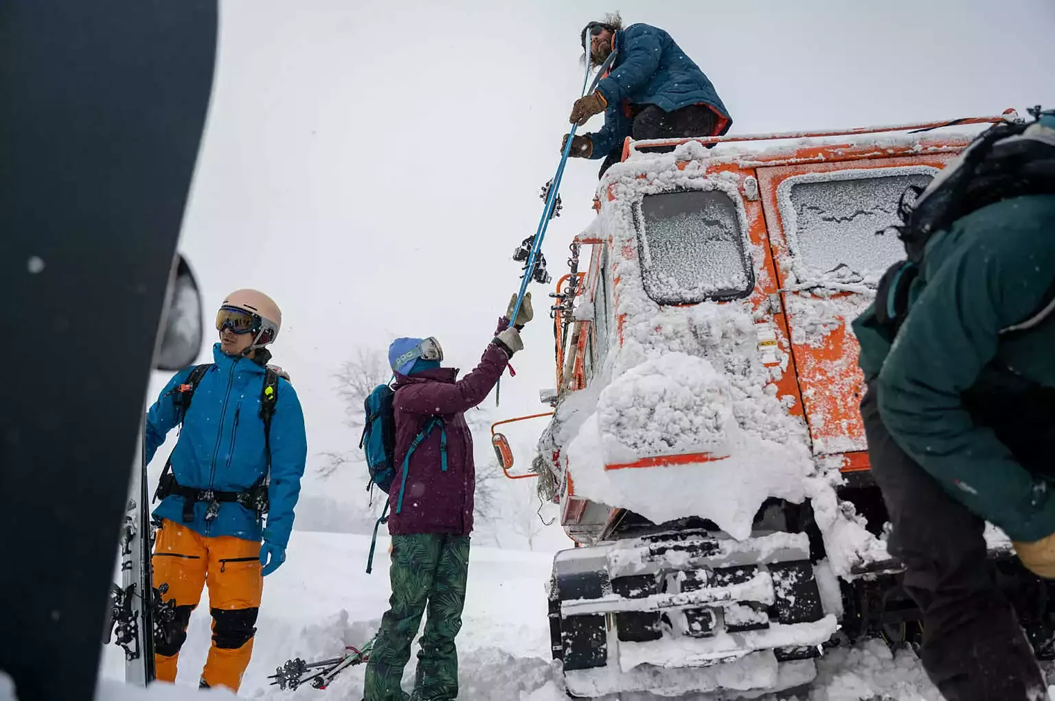 Group of skiers in snowy weather helping a person on a rescue stretcher near an orange rescue vehicle.