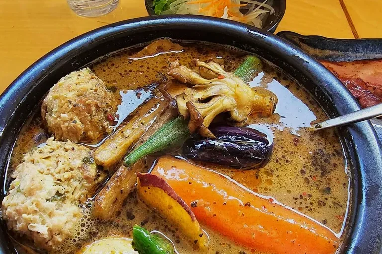 A bowl of soup with vegetables including carrots, okra, yellow squash, eggplant, and possibly some vegetable fritters or dumplings, with a side salad and a piece of bacon visible in the background.
