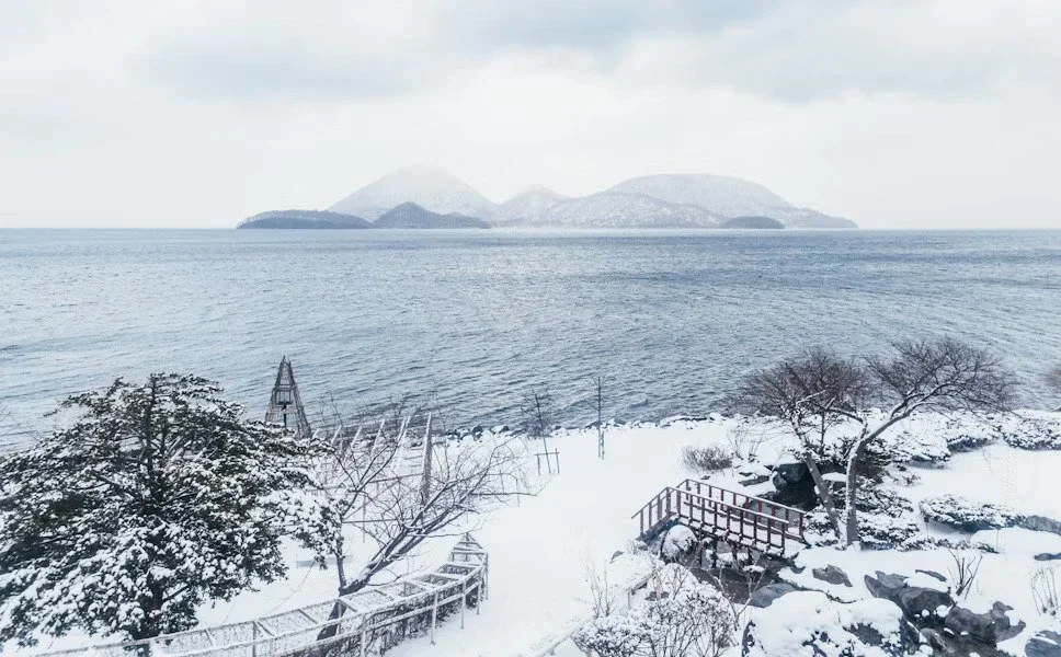 Snow-covered shoreline with trees, rocks, and a wooden deck, facing a large body of water with distant mountains or islands under a cloudy sky.