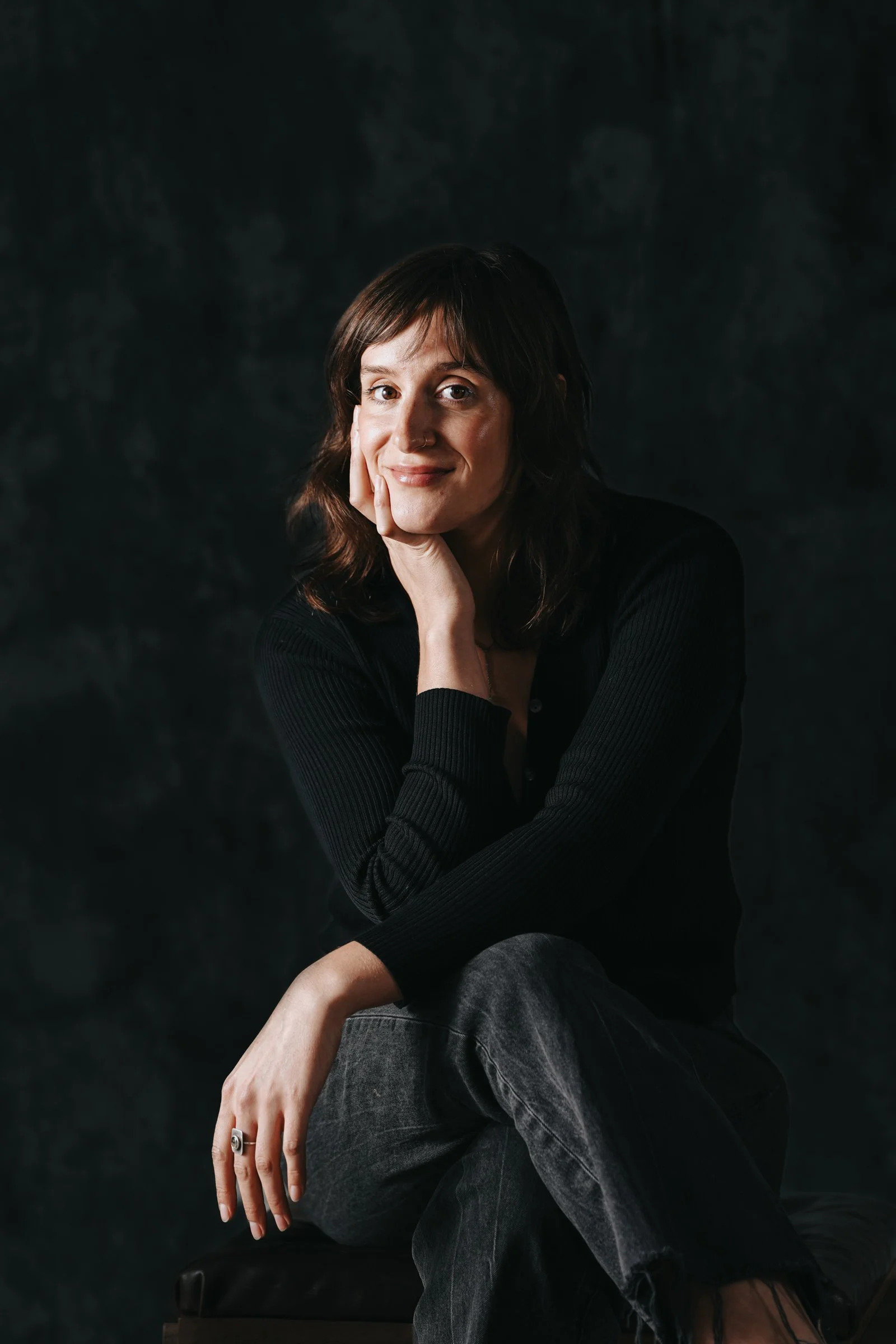 A woman with shoulder-length brown hair, dressed in a black long-sleeve top and gray jeans, sitting and resting her face on her hand, smiling softly against a dark background.