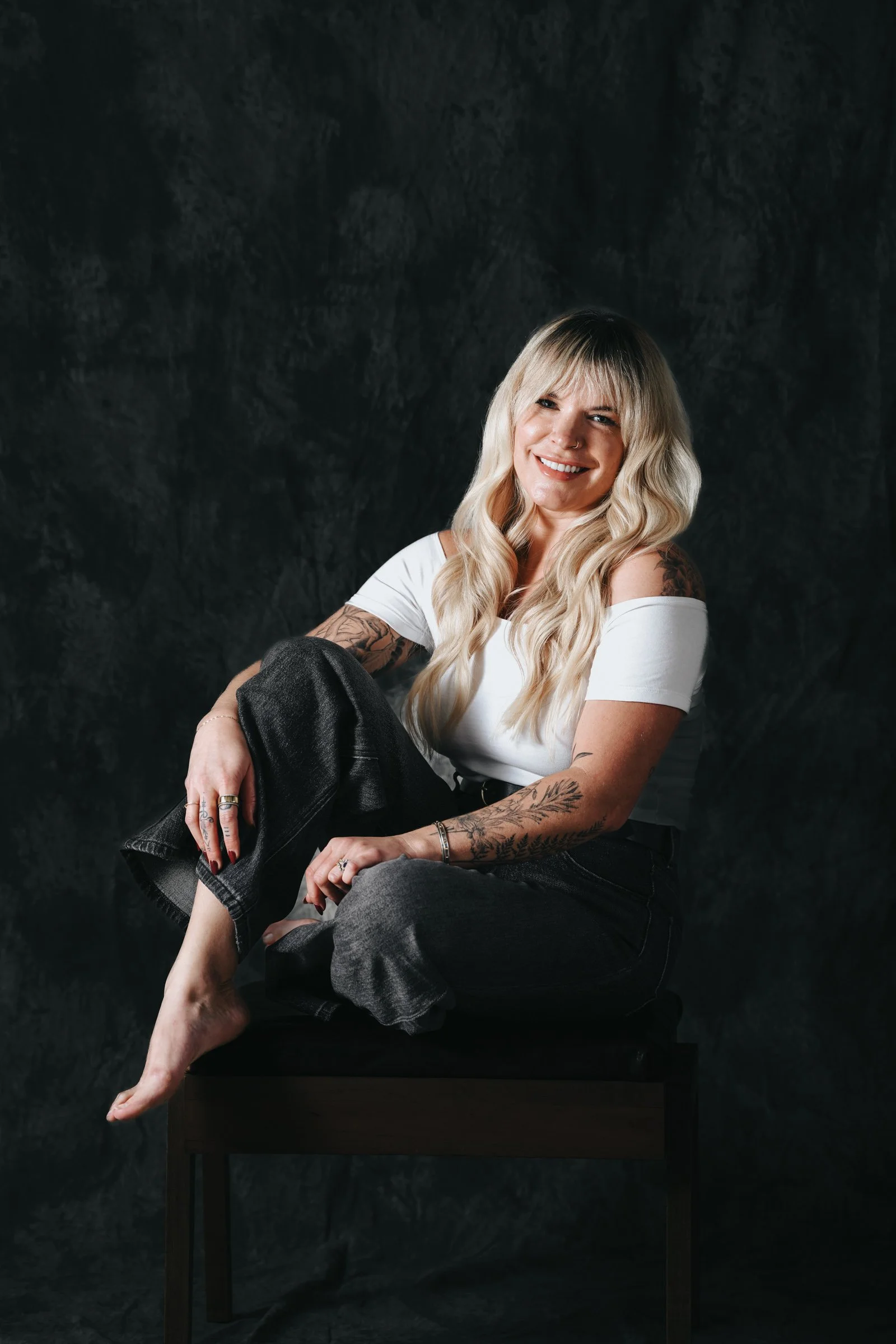 Smiling woman with long blonde hair, tattoos on arms, white off-shoulder top, black jeans, sitting on a small black stool against a dark textured background.