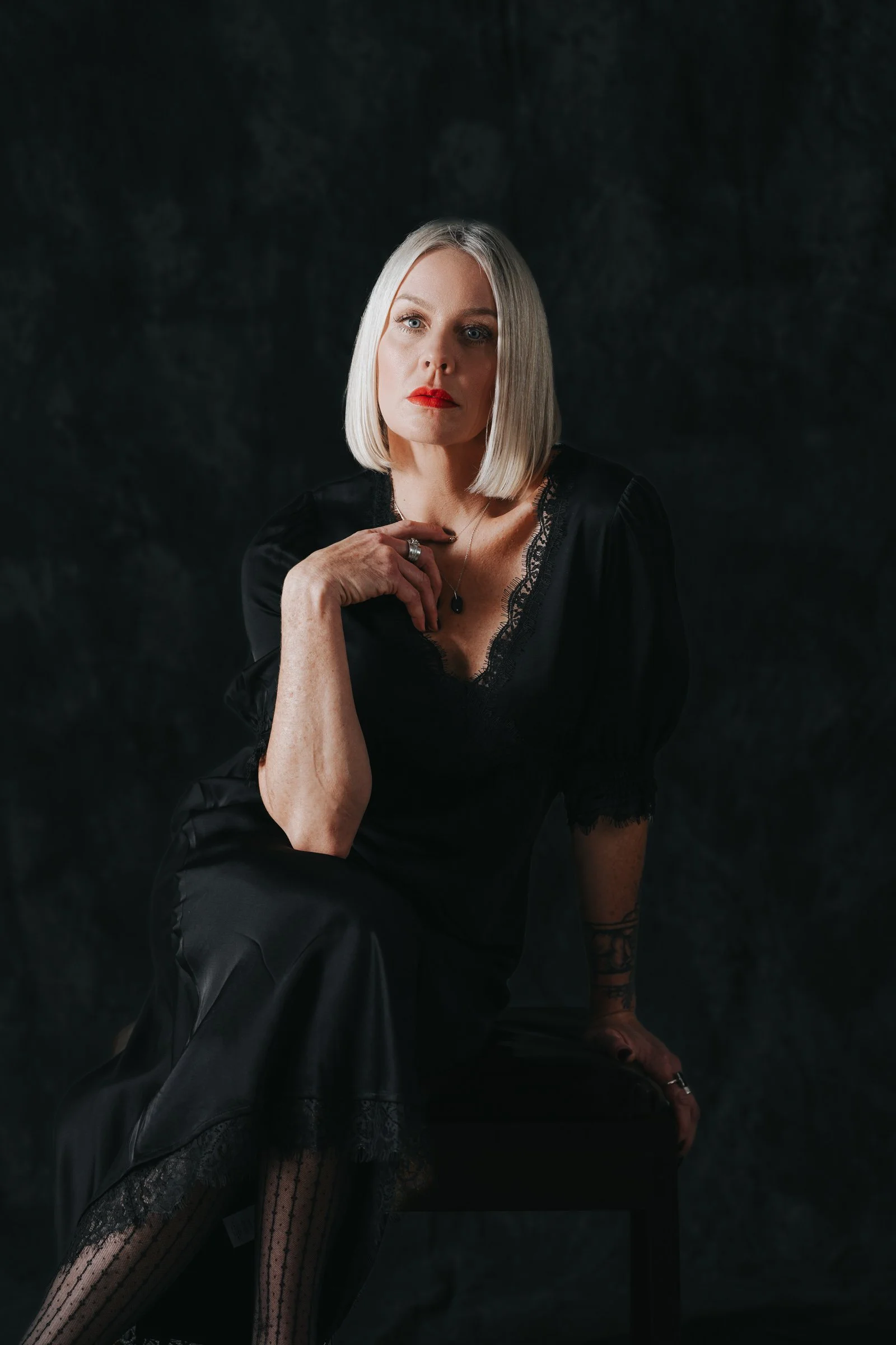 A woman with blonde hair, wearing a black lace-trimmed dress, seated on a chair in a studio with a dark background, looking at the camera.