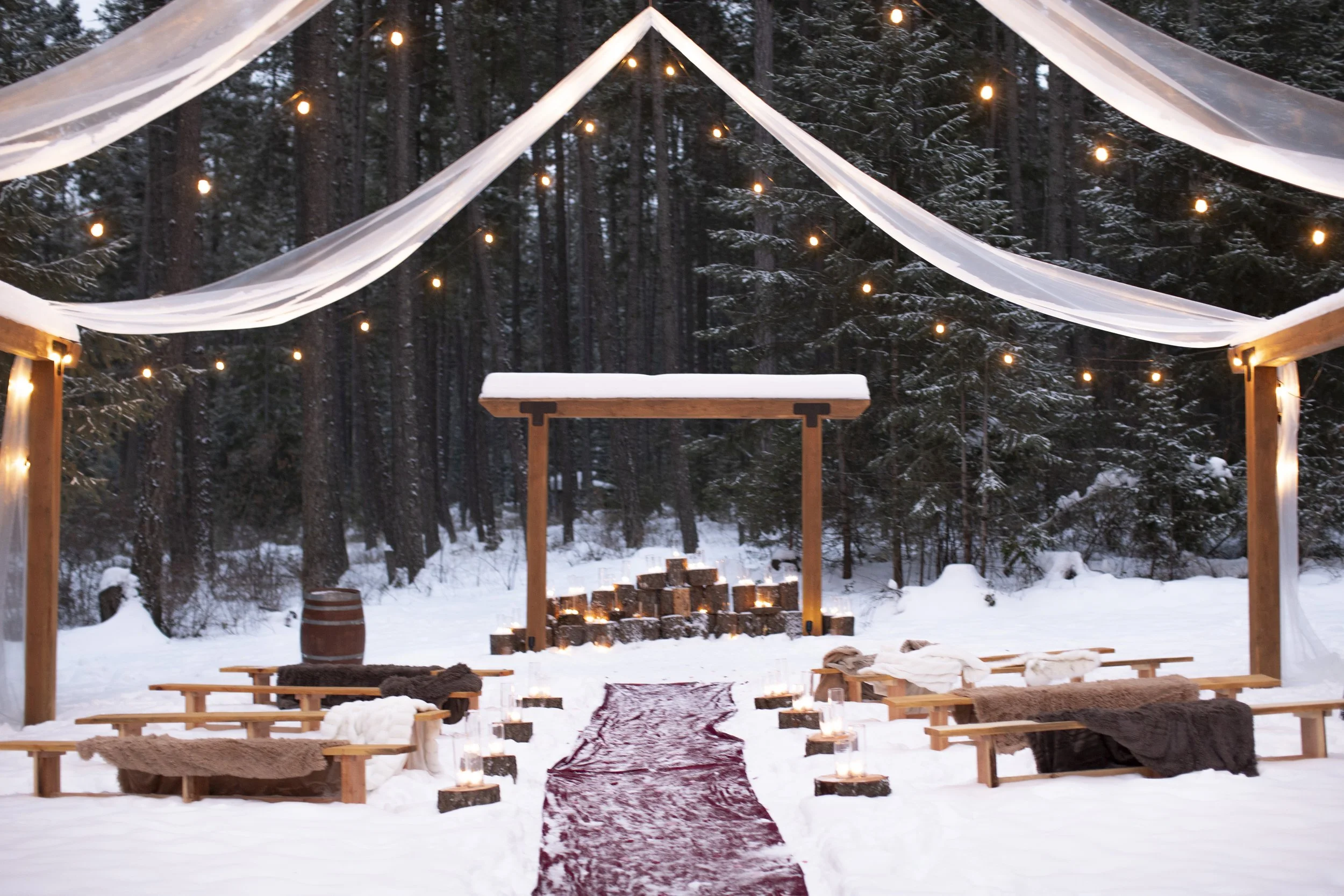 A cozy outdoor winter wedding setup in the snow with wooden benches, soft blankets, and candles, under a canopy with string lights and a forest background.