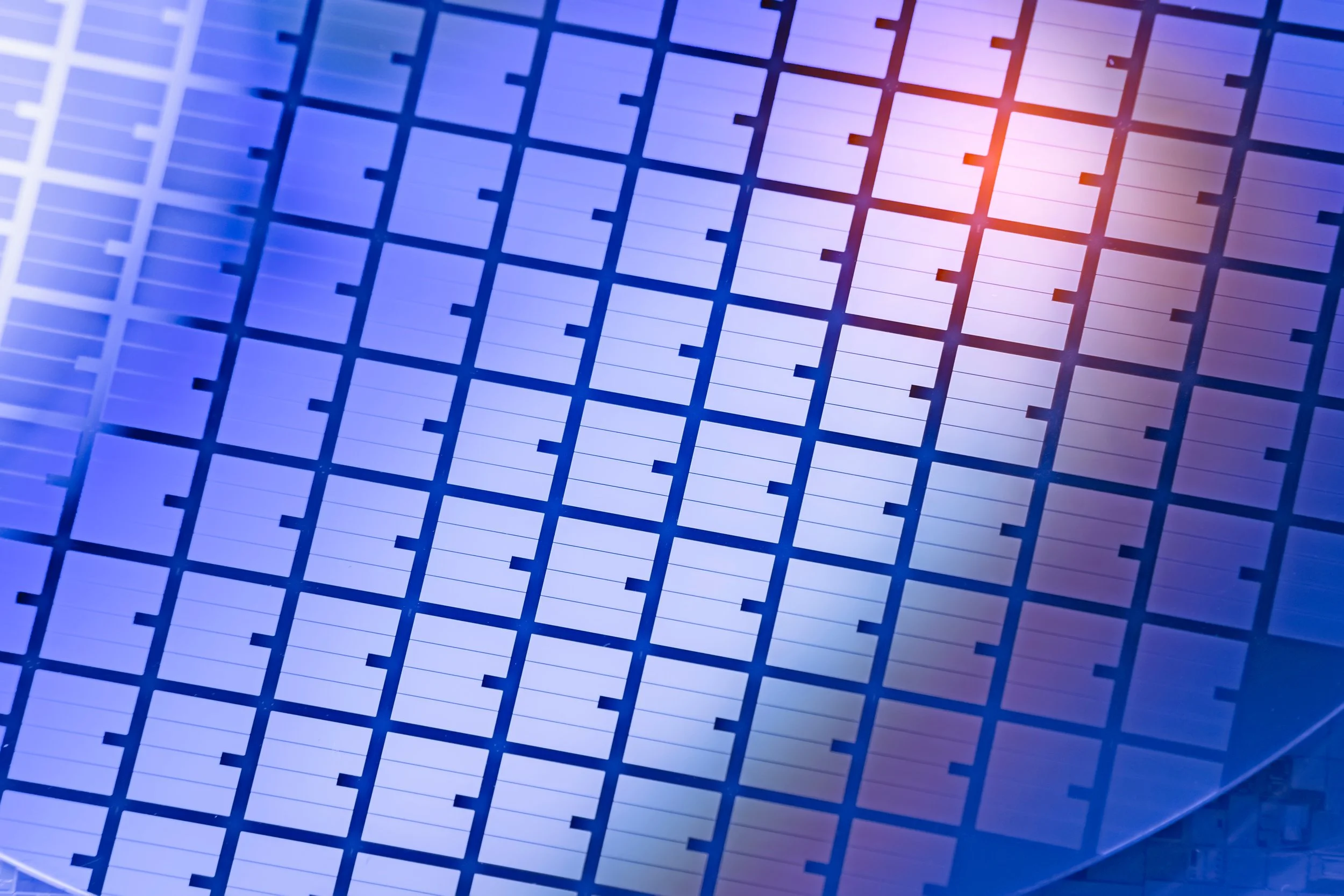 Close-up of a silicon wafer with a grid pattern used in semiconductor manufacturing, displaying a reflective surface with blue and purple hues.