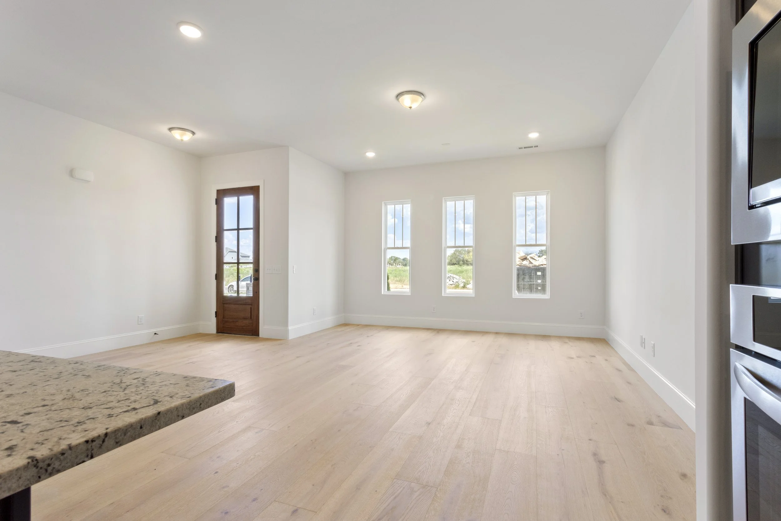 Empty living room with white walls, light wood flooring, three tall windows, a wooden door, and a view of sky and trees outside.