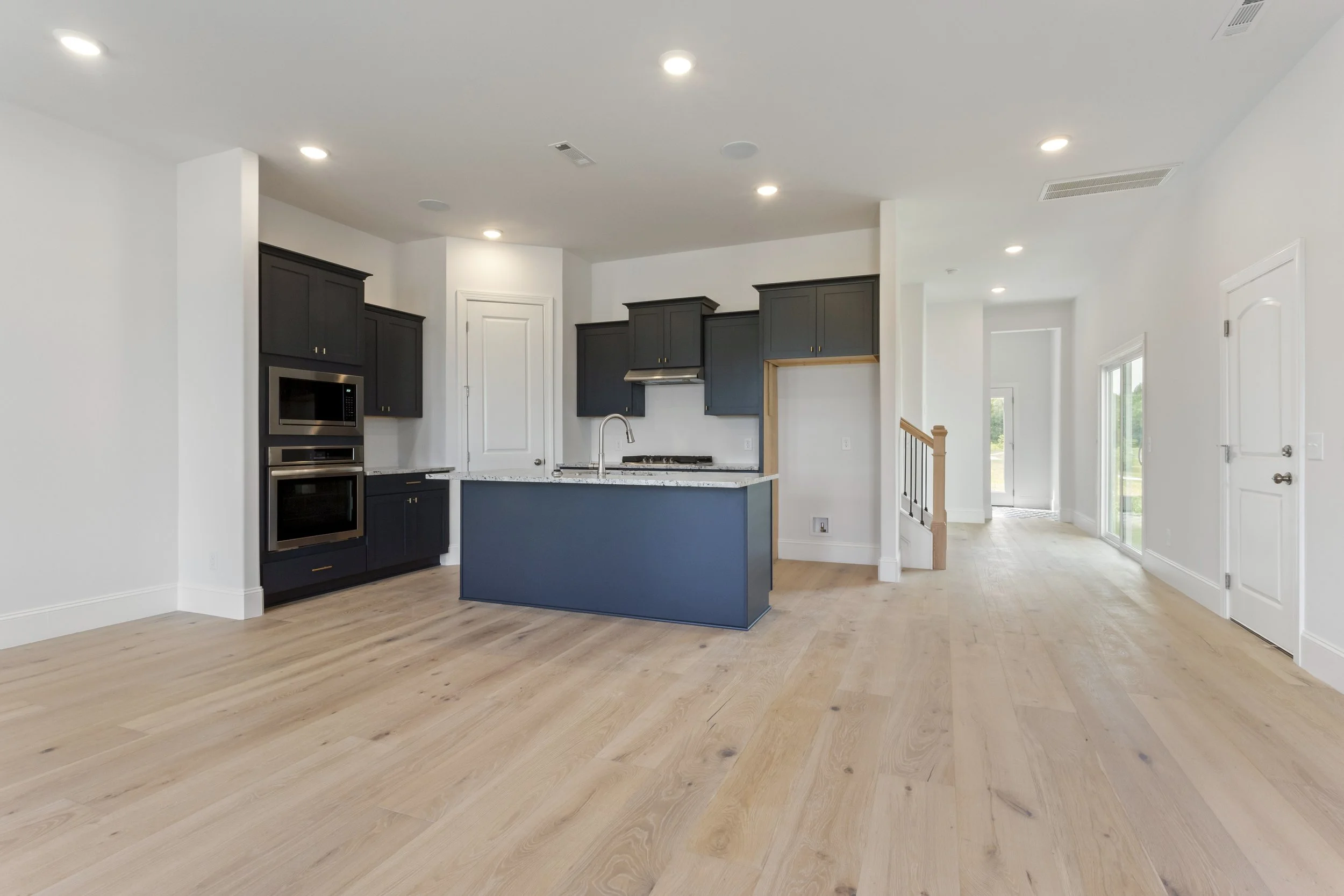 Open-concept modern kitchen with black cabinets, a blue kitchen island, stainless steel appliances, light hardwood floors, and white walls.