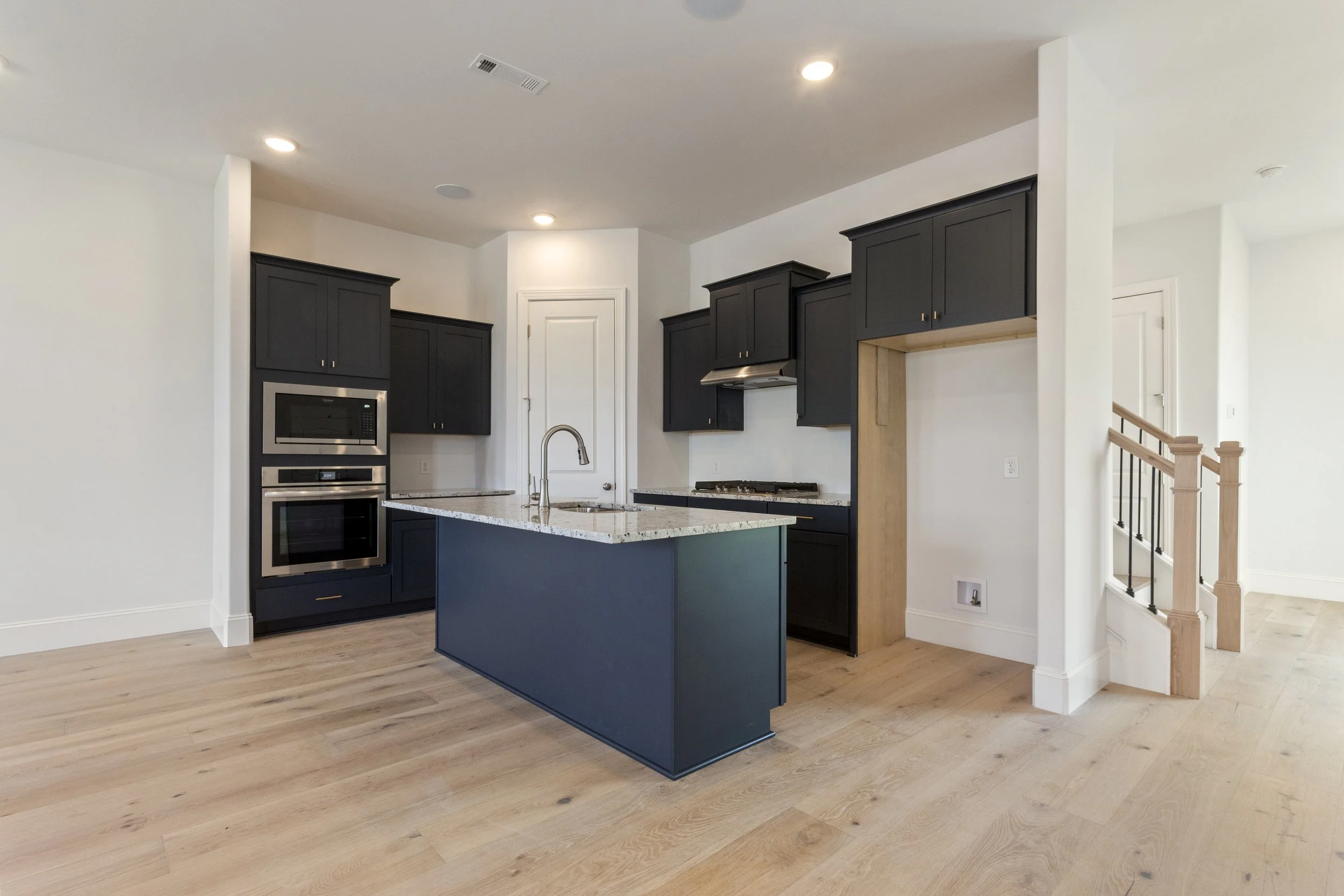 Modern kitchen with black cabinets, a central island with a granite countertop, stainless steel appliances, light wooden flooring, and a staircase on the right.