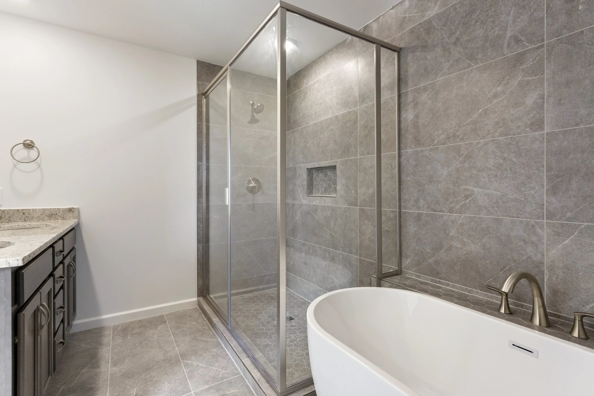 Modern bathroom featuring a glass shower enclosure, a white freestanding bathtub, dark wood vanity with granite countertop, and gray tiled walls and floor.