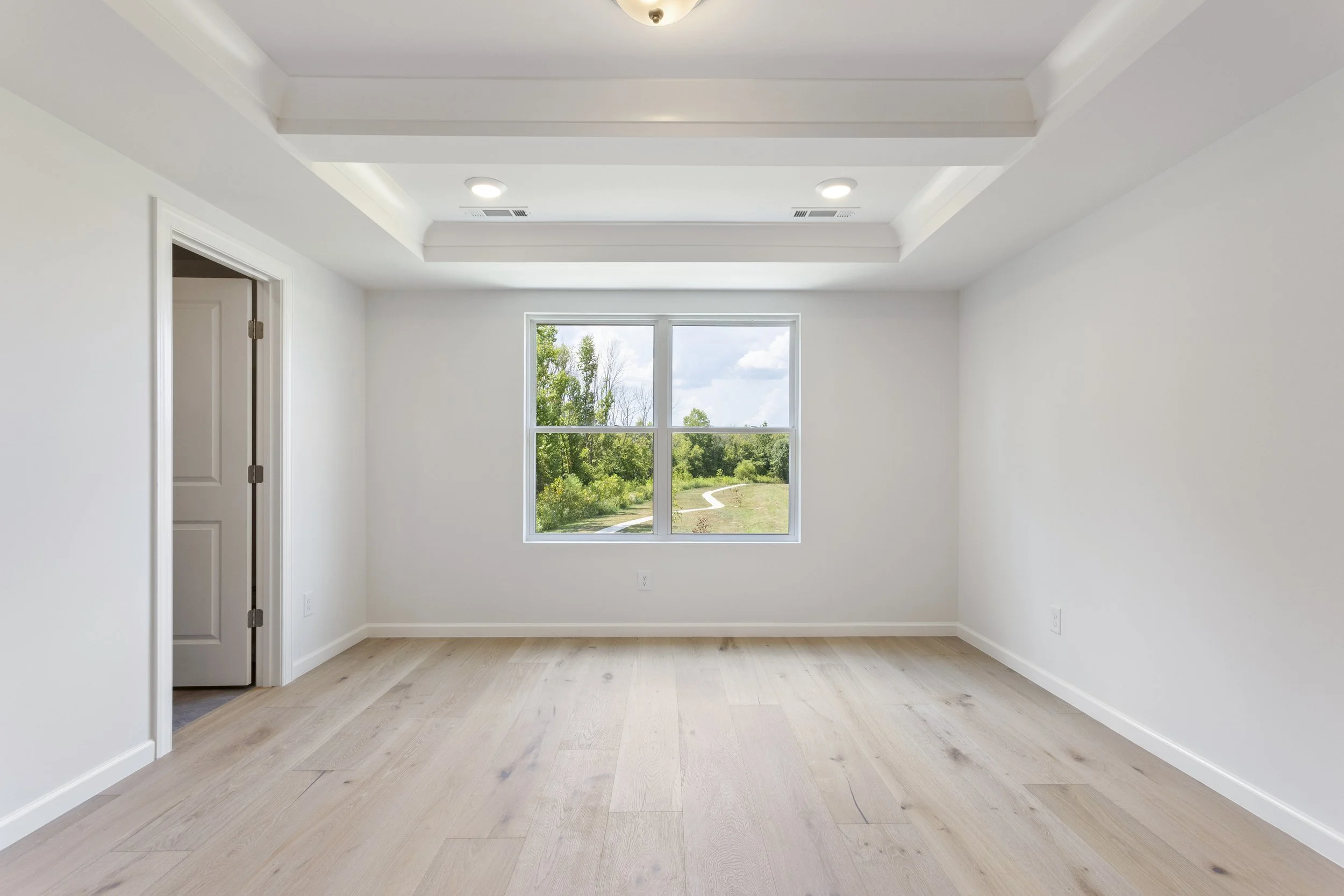 Empty room with white walls, light wood flooring, a window showing greenery outside, and a ceiling with recessed lighting.