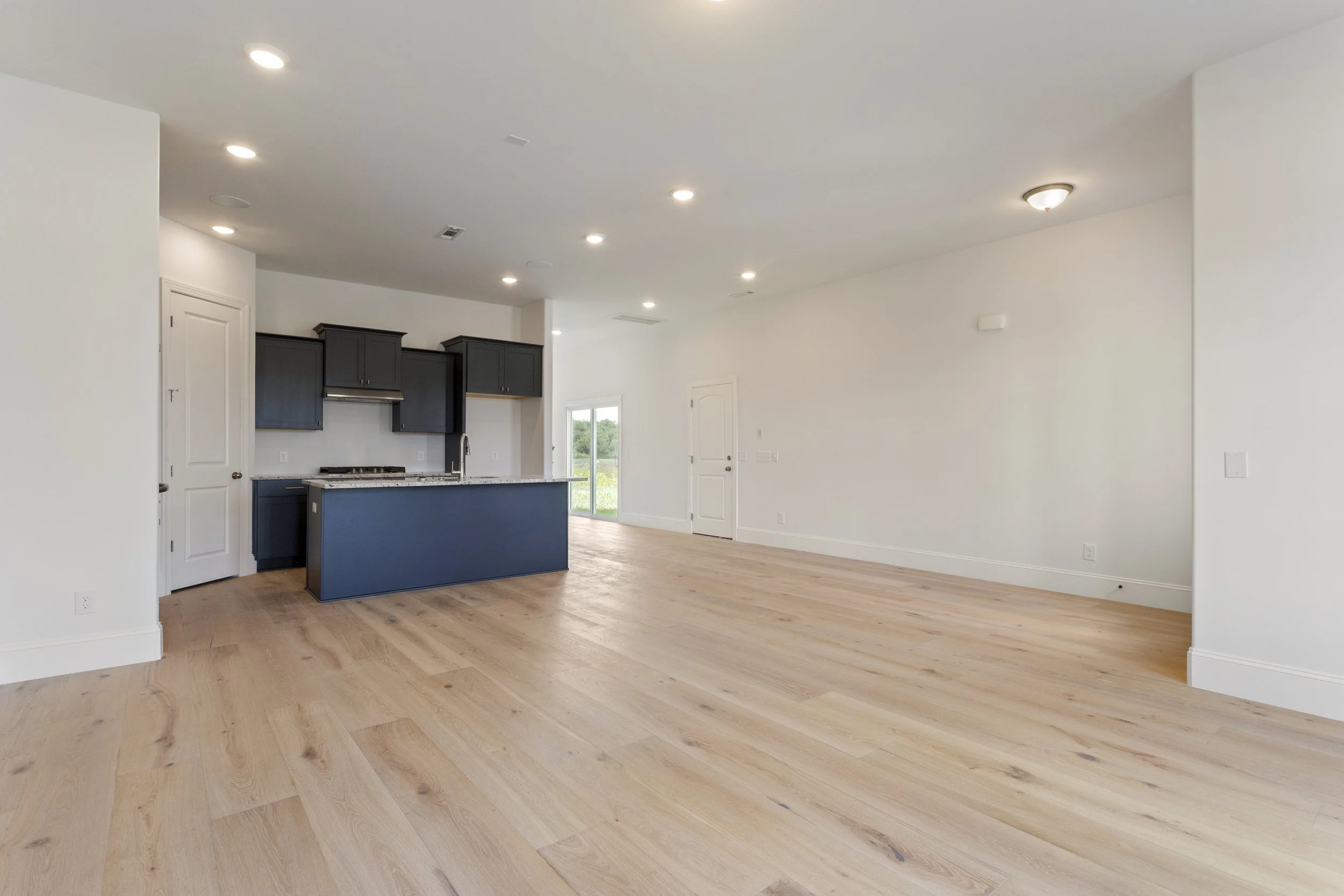 Empty modern open-concept living space with light wood flooring, a kitchen area with dark cabinets, and a door leading outside.
