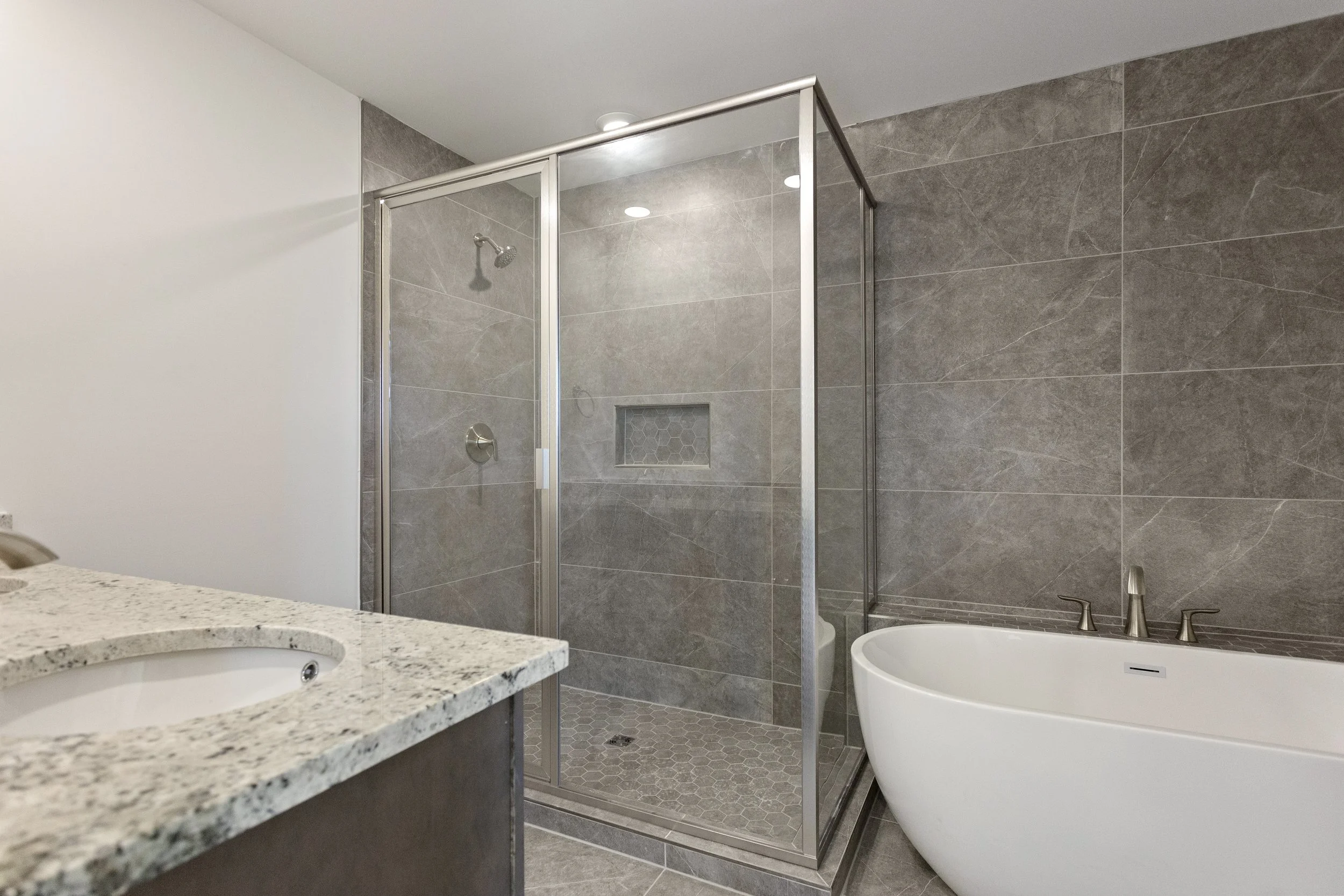 Modern bathroom with a corner shower with glass doors, gray tile walls, a built-in shower niche, a white freestanding bathtub, and a granite countertop bathroom sink.