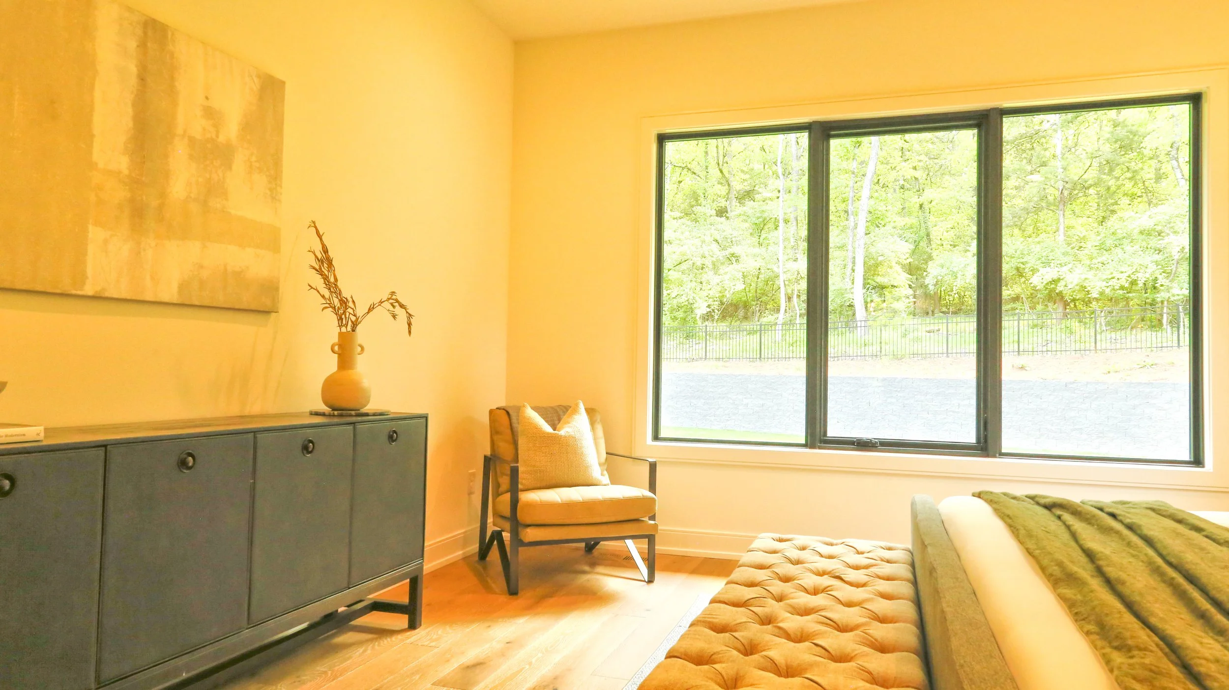 Living room with large window showing green trees outside, beige tufted ottoman, dark blue sideboard with a decorative vase and dried branches, and a beige armchair with a pillow.