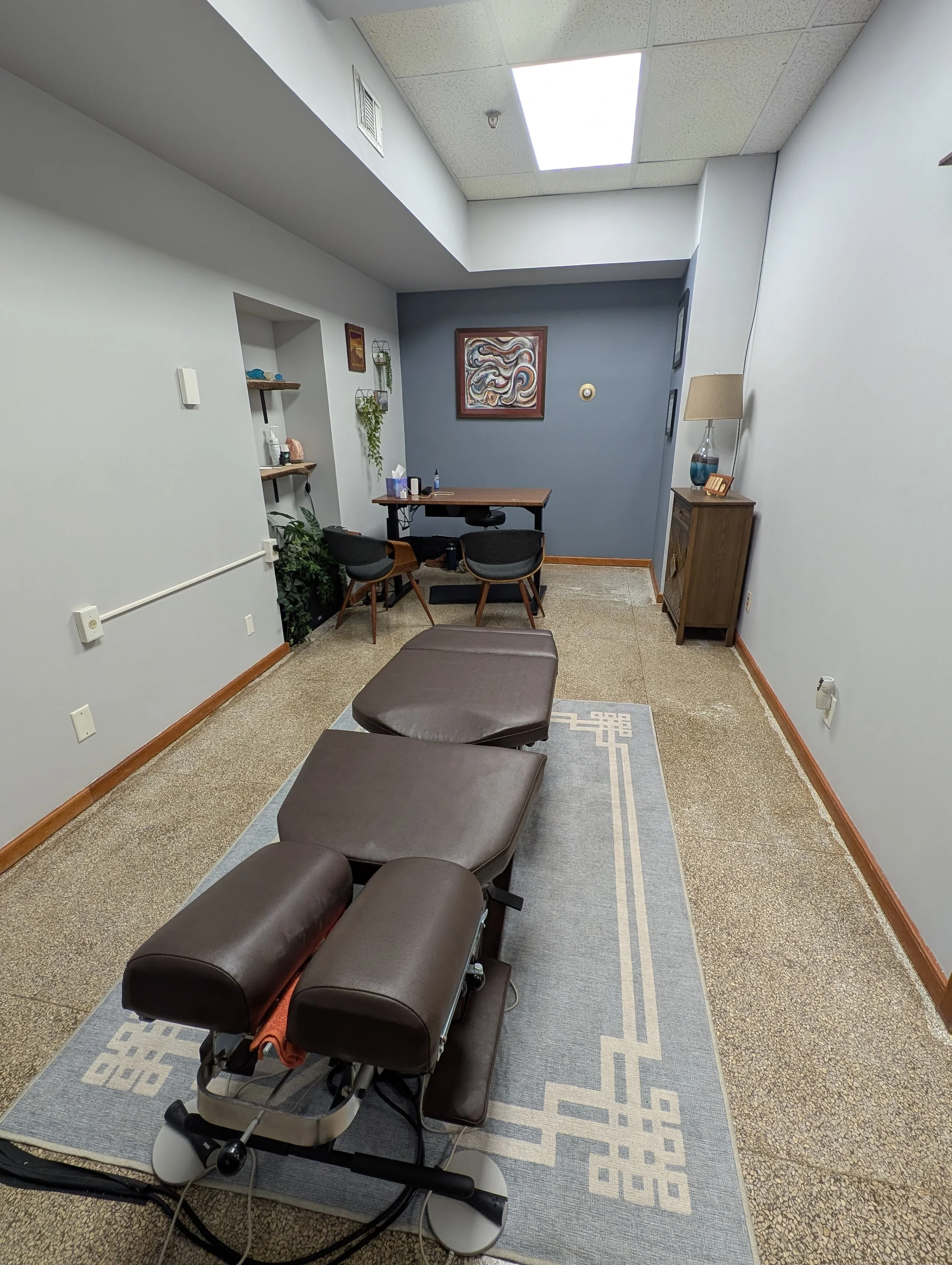 Treatment room with a chiropractic table, two chairs, a desk, and various decorations.