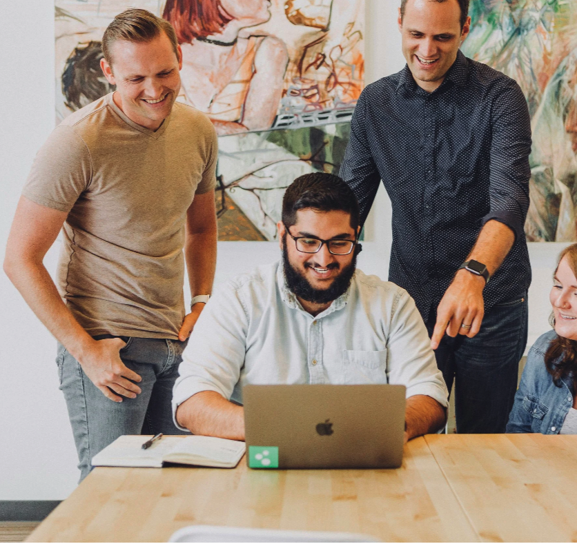 Four people gathered around a human first digital learning labs table, two standing and two sitting, looking at a laptop and smiling in an office setting.