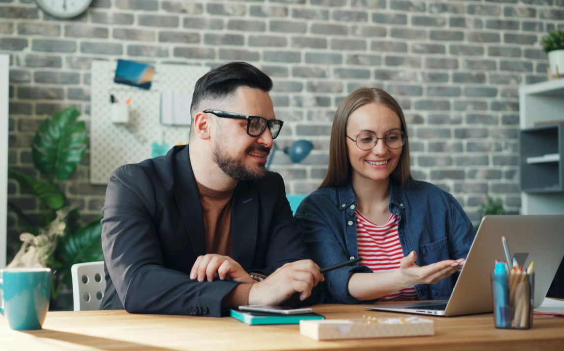Two coworkers, a man and a woman, look at a laptop screen together in an office with a brick wall background.
