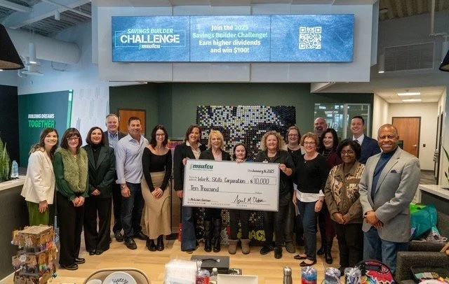 Group of people standing inside a modern office, holding a large check for ten thousand dollars, with a digital screen above displaying a savings challenge announcement.