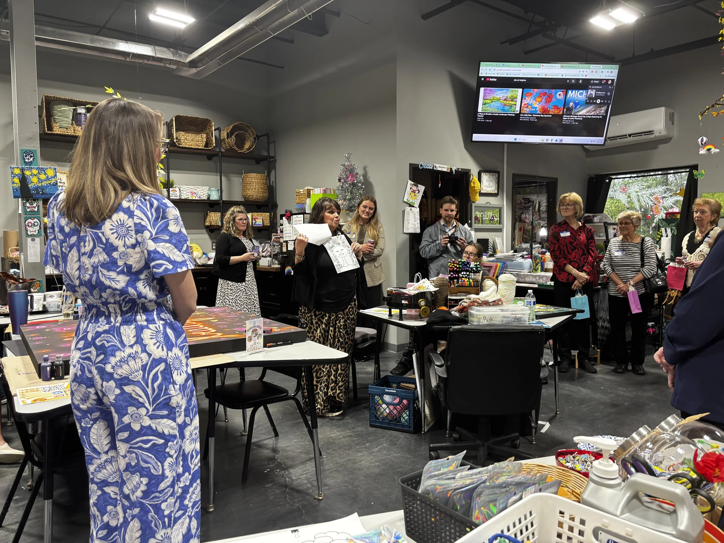 A group of people gathered in an indoor craft or art store, with shelves filled with baskets and art supplies, and a decorated Christmas tree in the background. Some individuals are holding artwork, a woman appears to be speaking, and others are watching or taking photos.