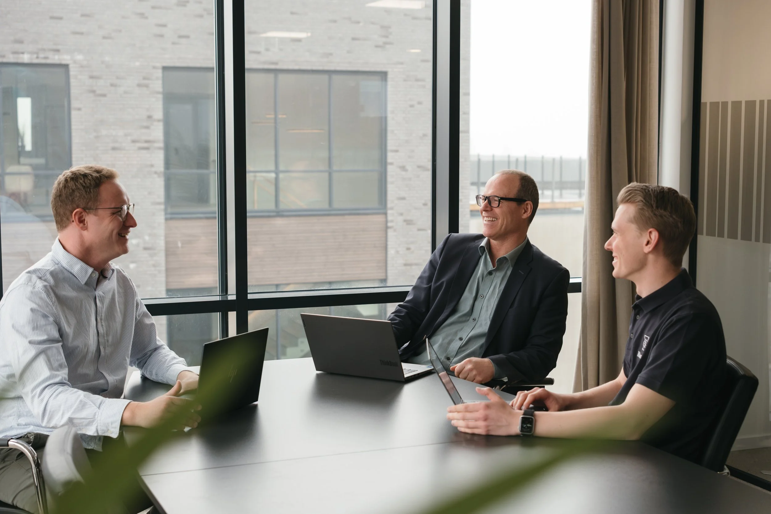 Tre personer i et møterom som smiler og snakker, alle med laptop foran seg, store vinduer bak dem.
