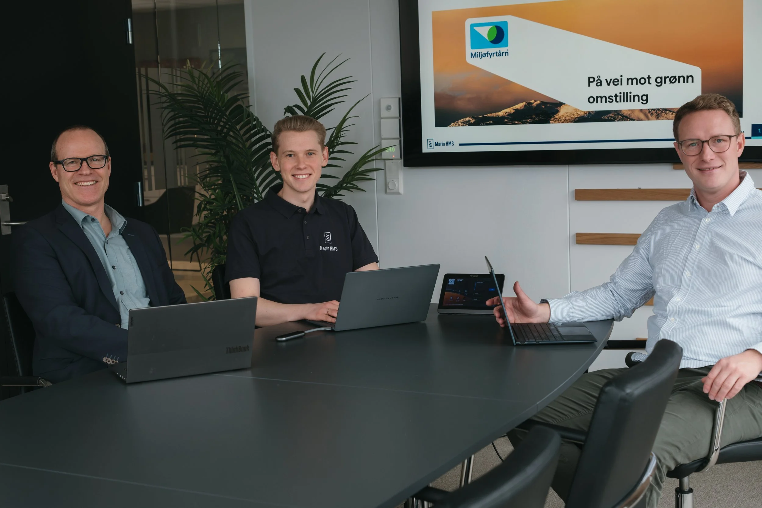 Tre menn sitter rundt et konferansbord med bærbare datamaskiner, foran en skjerm med teksten 'På vei mot grønn omstilling' og en logo. De smiler og ser fornøyde ut i et lyst møterom med planter.