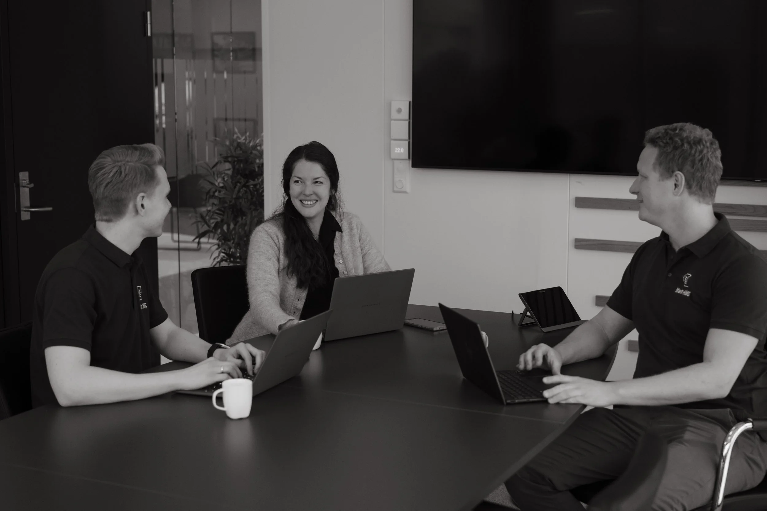 Tre personer foran en konferansbord, to menn og en kvinne, som smiler og har laptopper i et møterom.