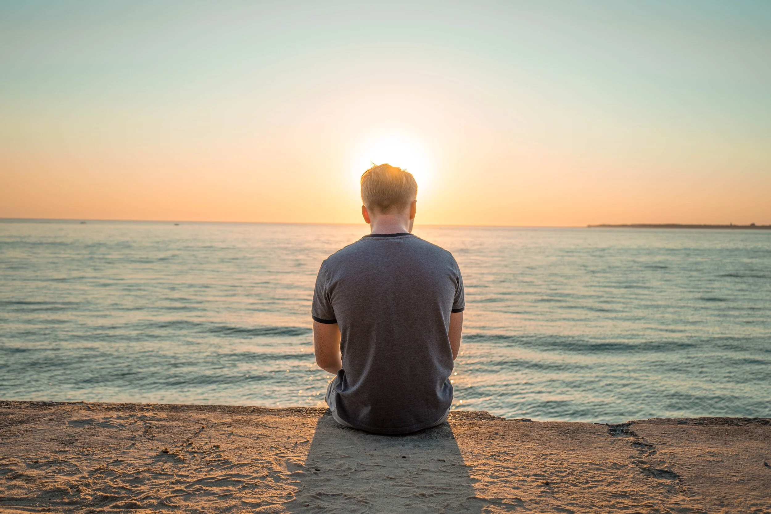 Person sitting on a concrete surface at the beach during sunset, facing the ocean with the sun near the horizon.