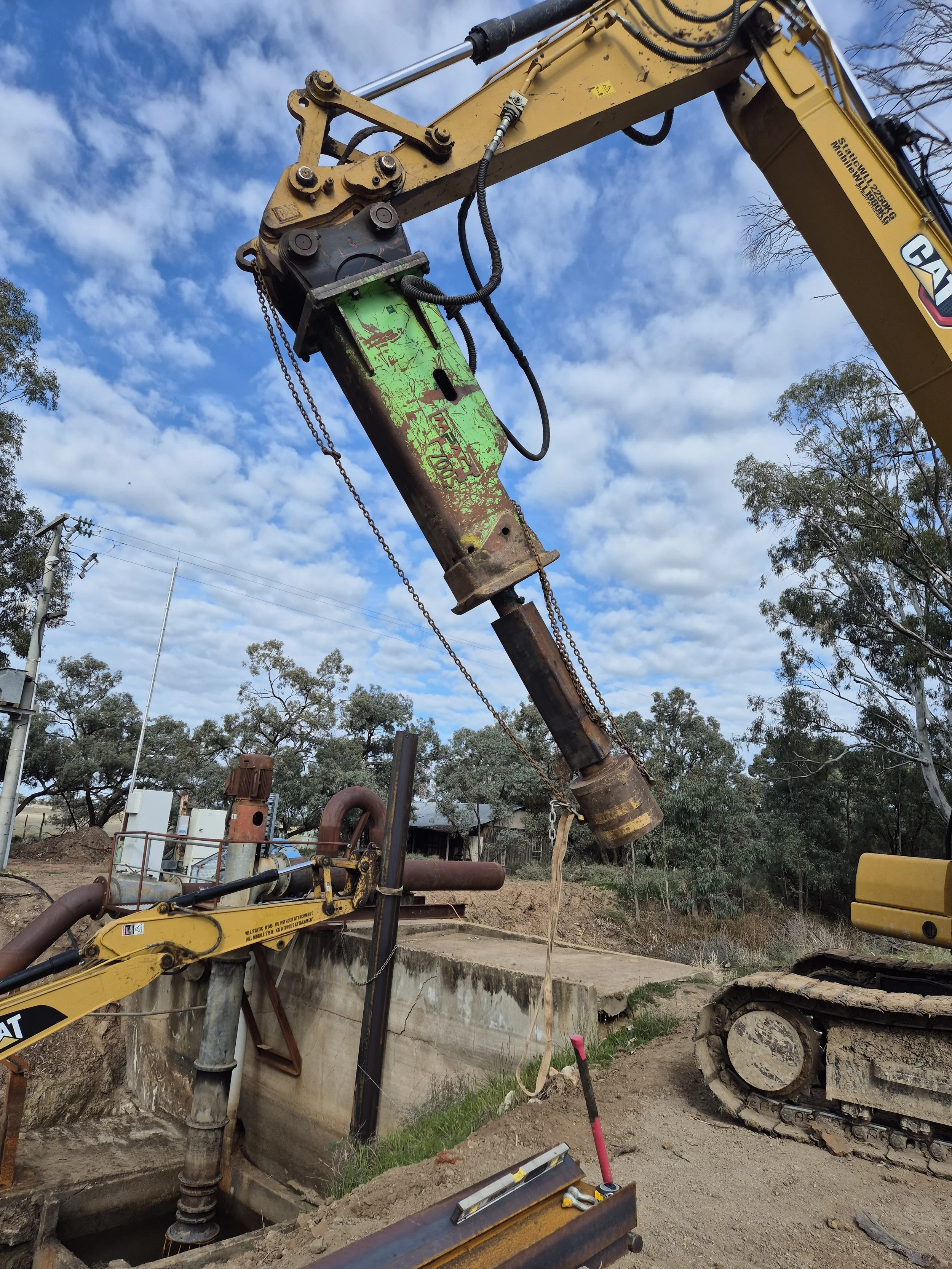 Construction site with a large yellow hydraulic excavator and a steel casing pipe being installed into the ground; blue sky with scattered clouds, trees in the background.