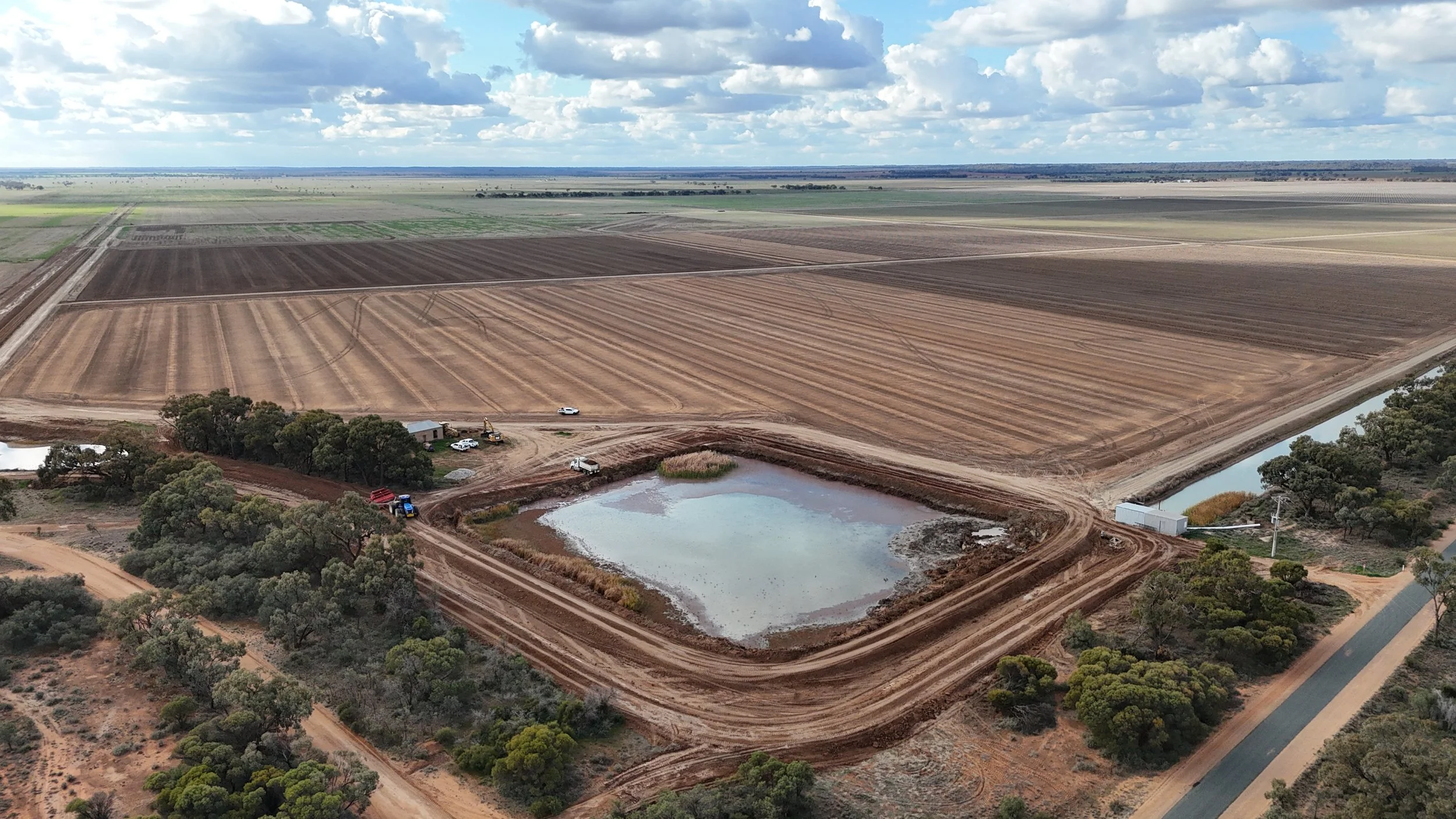 Aerial view of a farm with large, rectangular fields, a pond, and a dirt road, under a sky with clouds.