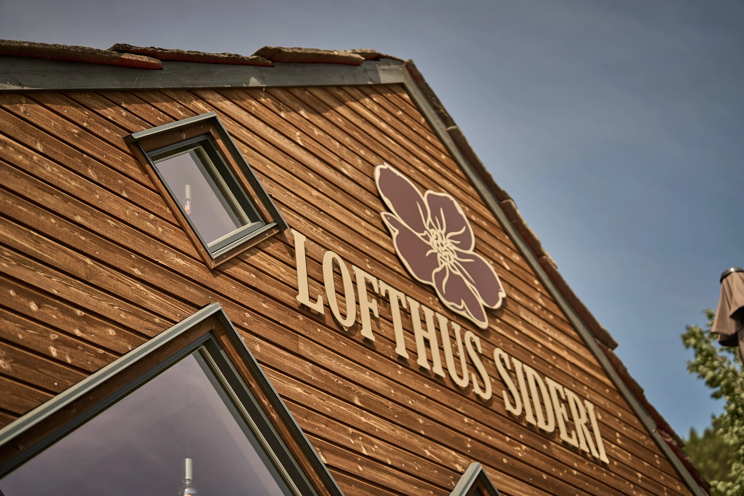 Close-up of a wooden building with a sign that reads 'LOFT'. There are two small, angled windows on the building's exterior and a large flower logo next to the sign. The corner of the roof is visible under a clear sky.