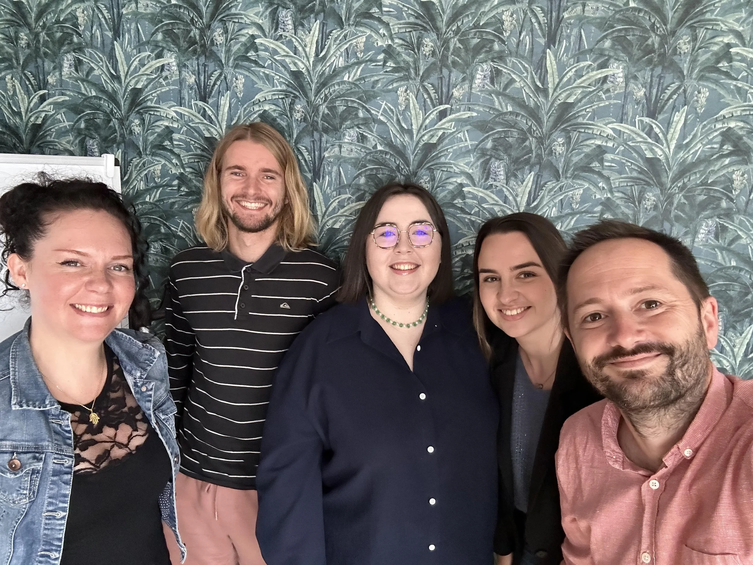 Groupe de cinq personnes souriantes posant ensemble devant un mur à motifs végétaux.