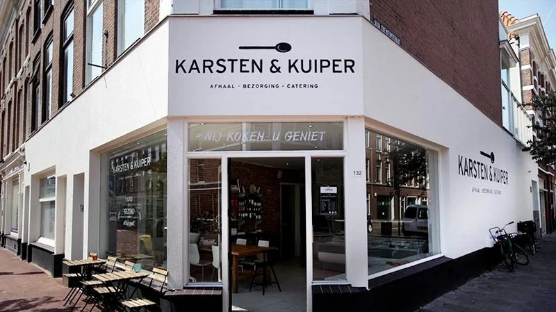 Winkelpand met de naam Karsten & Kuiper, gespecialiseerd in afhaal, bezorging en catering, met grote ramen en een zitgedeelte buiten.