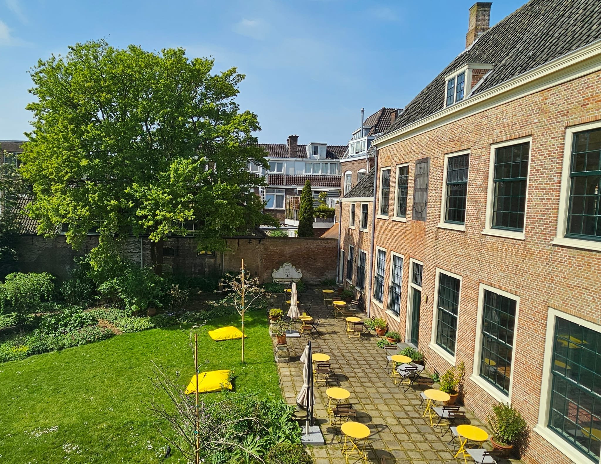 Een binnenplaats met een grasveld, tuinmeubilair met gele en beige tafels, parasols, en een grote groene boom, omgeven door oude bakstenen gebouwen onder een blauwe lucht.