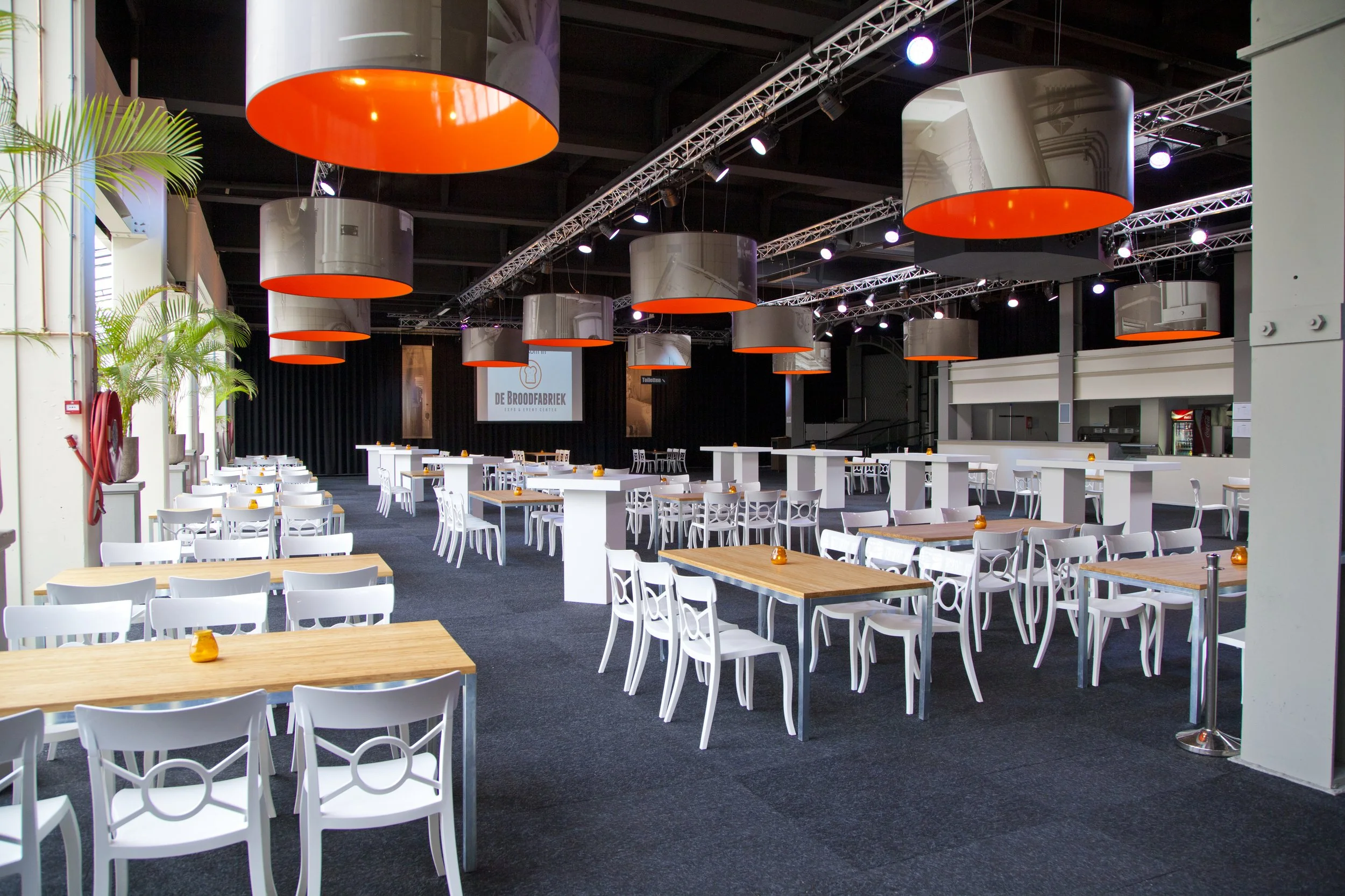 Expositieruimte met witte stoelen, houten tafels en grote ronde lichtarmaturen met oranje binnenkant, een bar of buffet aan de rechterkant, en een groot scherm met logo 'De Brooodfabriek' aan de achtergrond.