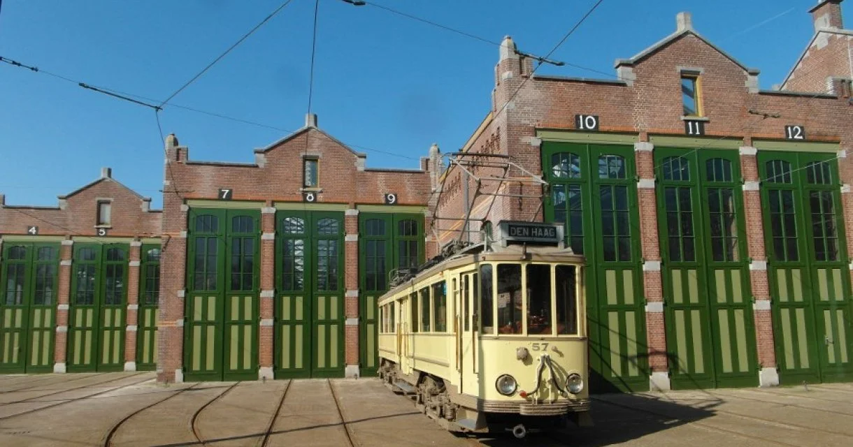 Een tram met nummer 57 rijdt in de stad Den Haag, voor een gebouw met grote groene hangende deuren en een blauwe hemel.
